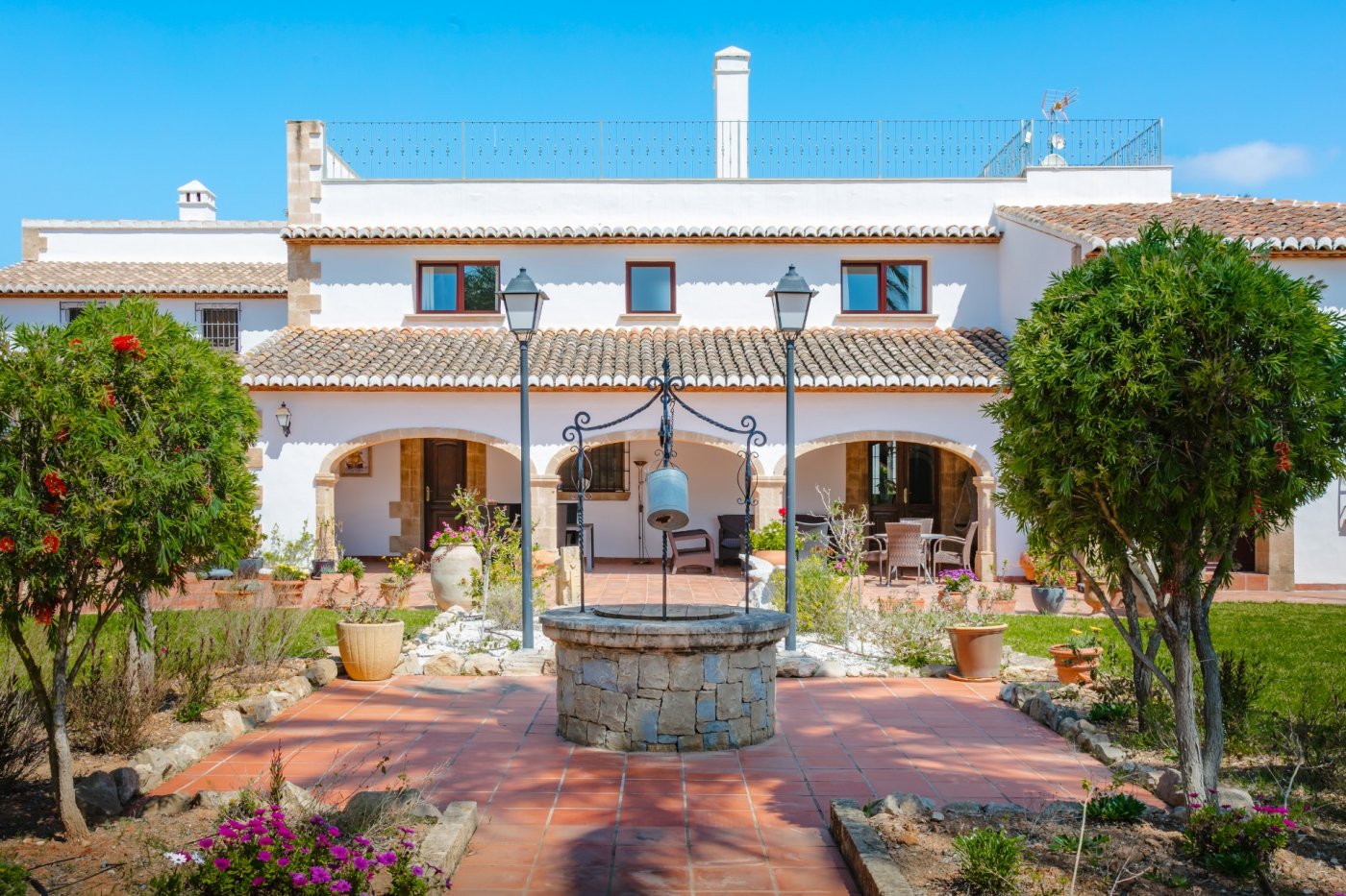 Patio exterior con pozo de piedra tradicional, suelos de terracota y fachada blanca con arcos de piedra tosca bajo un cielo despejado.