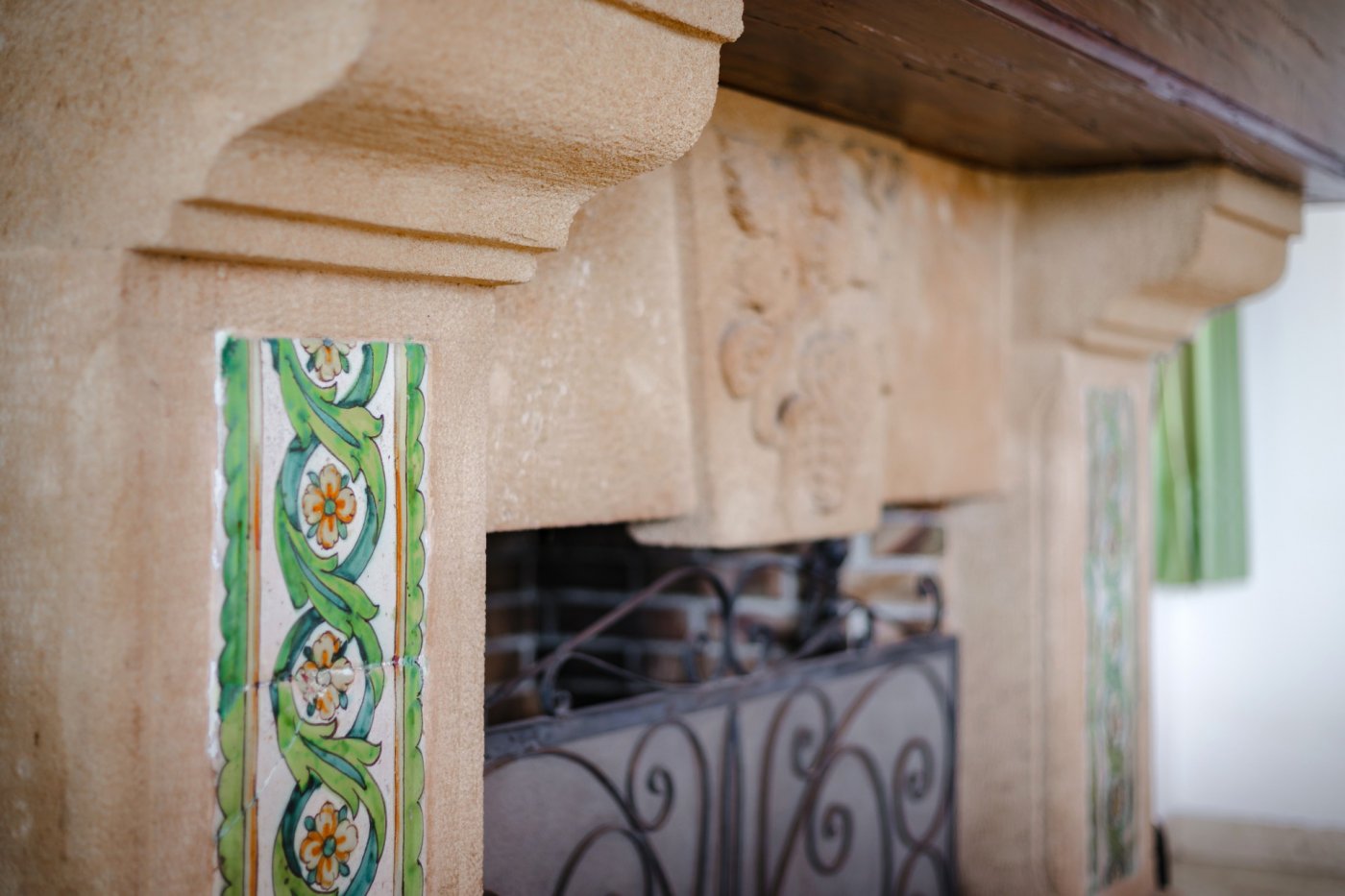 Primer plano de una chimenea de piedra tosca con azulejos cerámicos pintados a mano, talla ornamental central y rejilla de hierro forjado.
