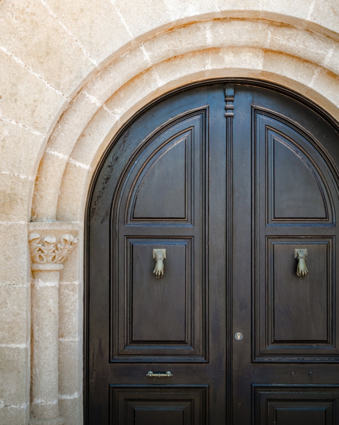 Puerta principal de madera oscura con arco de medio punto, enmarcada por piedra tosca y una columna decorativa con capitel tallado.