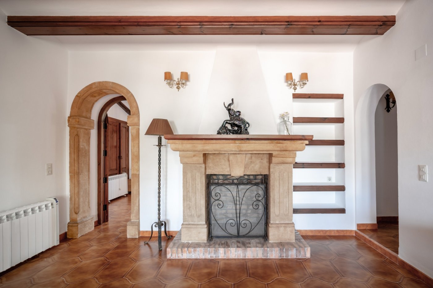 Salón con chimenea de piedra tosca labrada, vigas de madera vista y arco tradicional. El suelo de terracota complementa las estanterías de obra y la iluminación clásica.