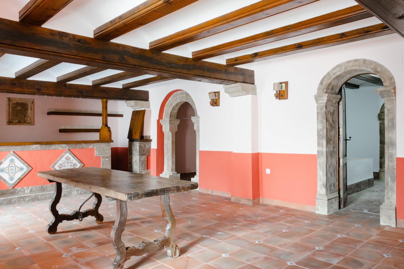 Salón rústico con vigas de madera a la vista, suelos de terracota y arcos de piedra tosca tallada. Incluye una mesa de madera maciza y detalles de azulejos tradicionales.