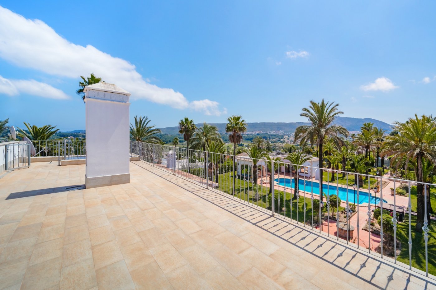 Terraza superior con suelo de gres cerámico y barandilla blanca, ofreciendo vistas panorámicas al extenso jardín con palmeras, la piscina privada y el valle de Jávea.