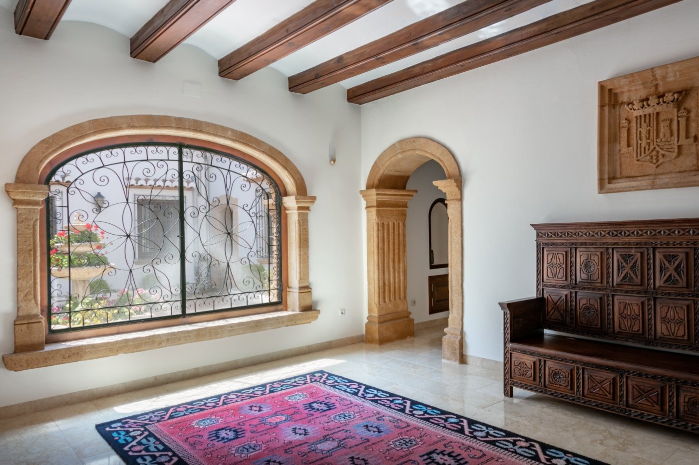 Vestíbulo con arcos de piedra tosca, vigas de madera y gran ventanal con rejería de forja que ofrece vistas al patio interior con fuente.