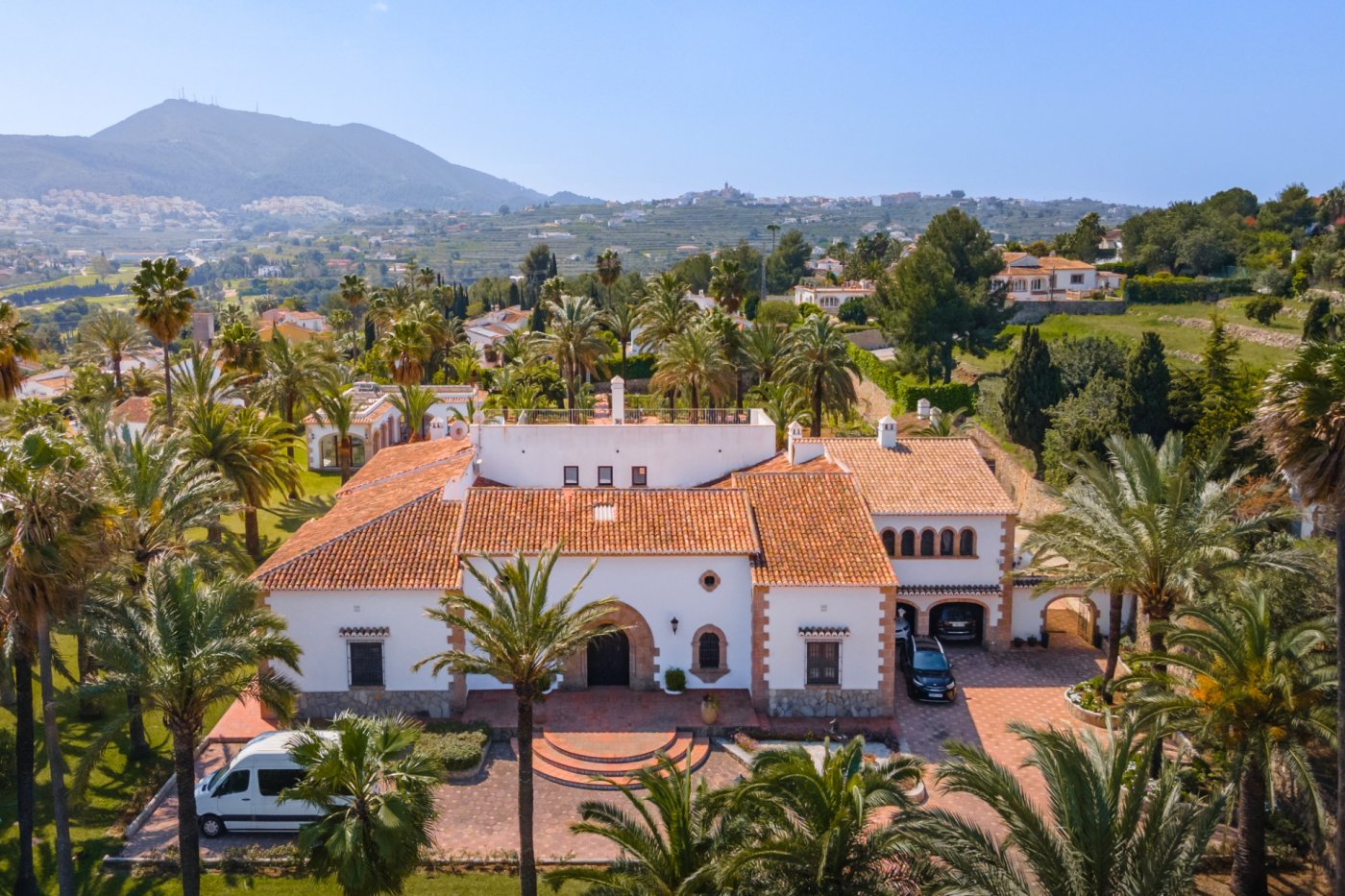 Vista aérea de la propiedad de estilo mediterráneo con fachadas blancas, arcos de piedra tosca y tejados de teja, rodeada de un extenso palmeral y vistas al valle.