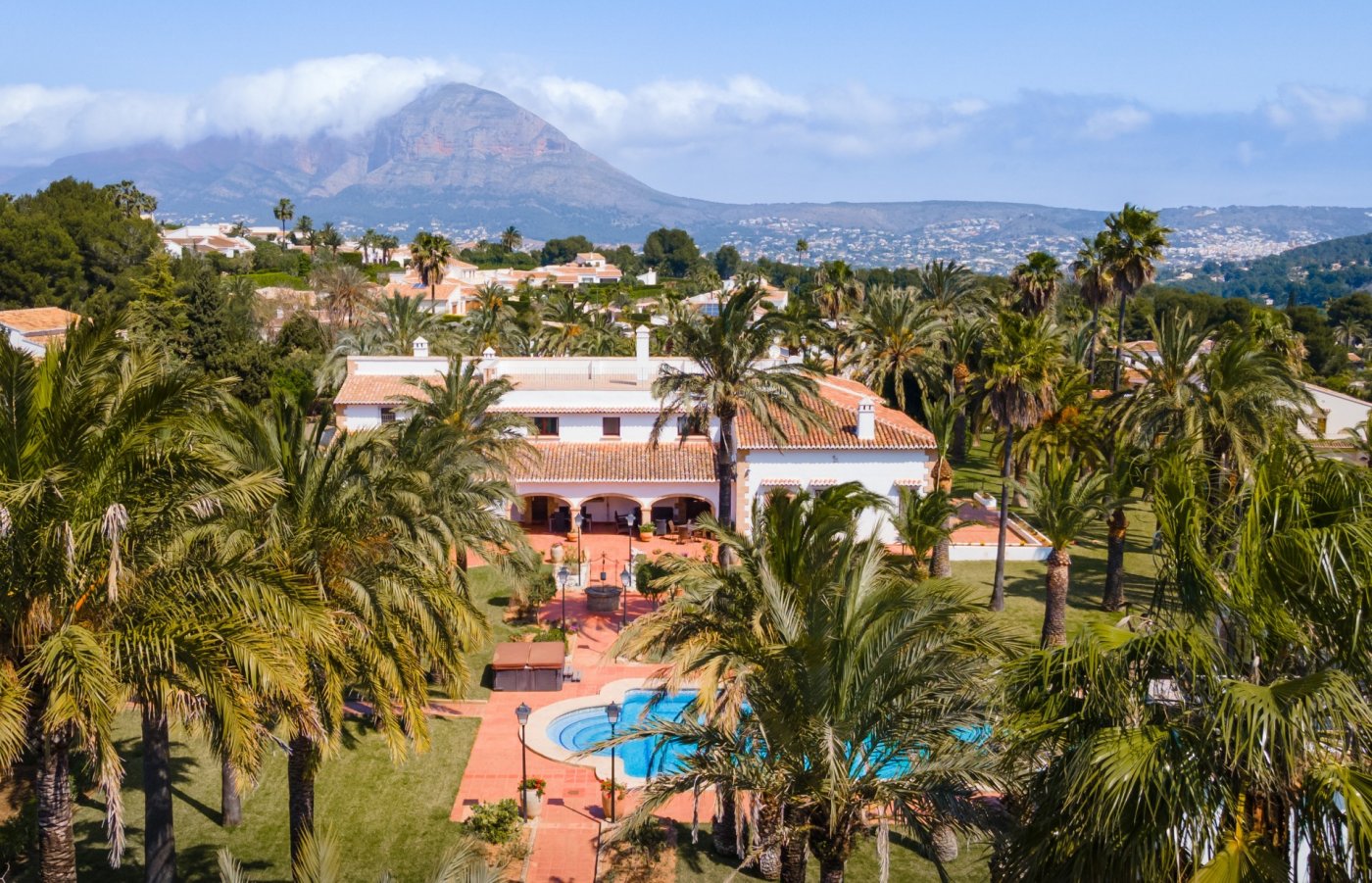 Vista aérea de la villa mediterránea con piscina, rodeada de un extenso palmeral y con vistas panorámicas al monte Montgó en Jávea.
