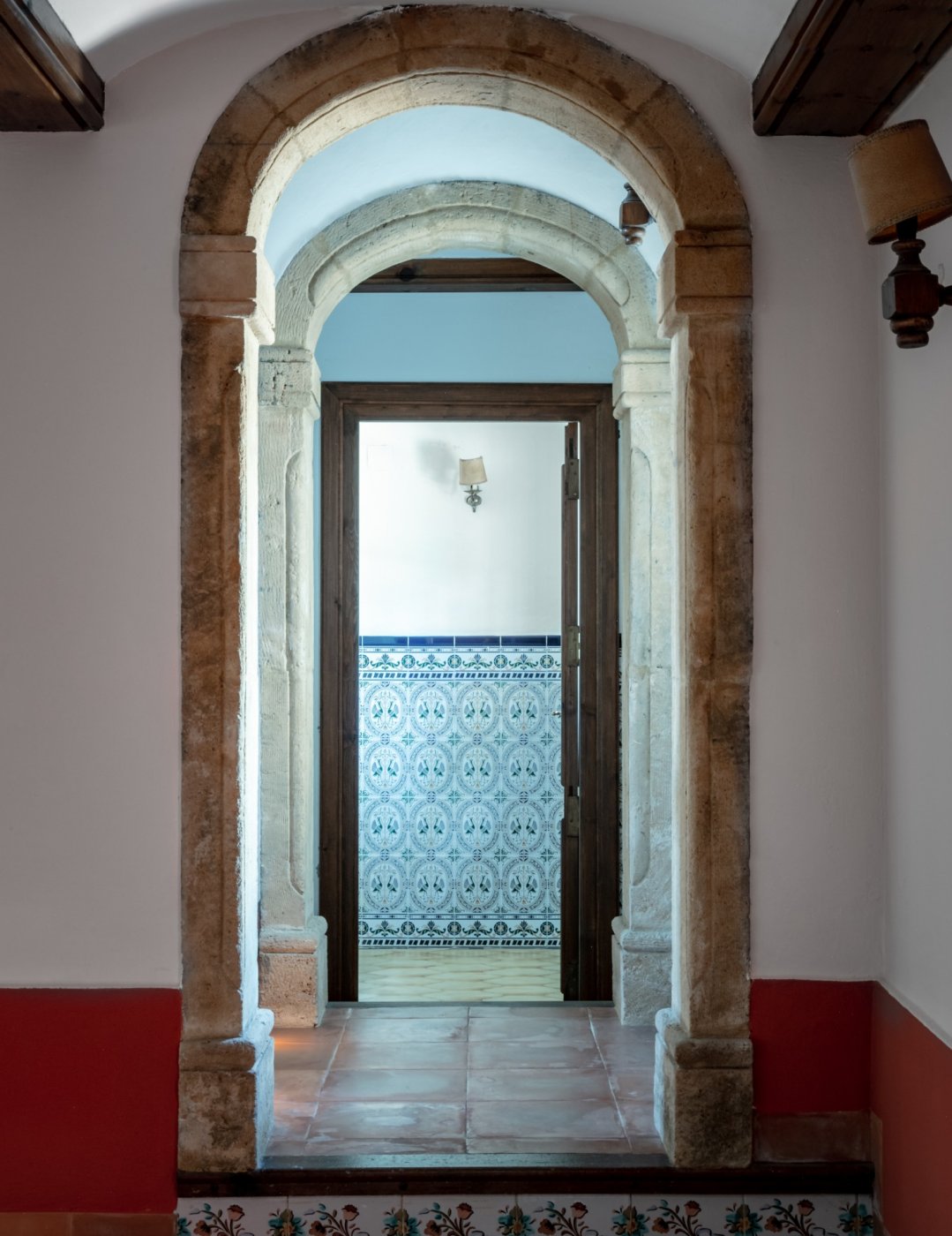 Vista de pasillo con arcos de piedra tosca natural que conducen a una estancia con azulejos decorativos tradicionales y suelos de barro cocido.