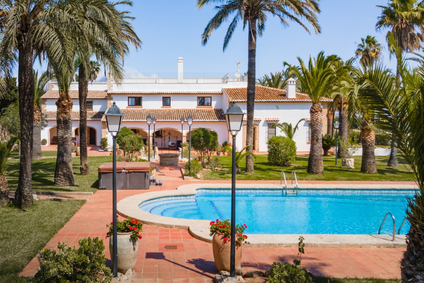 Vista exterior de la piscina de 16x7m y el jardín con palmeras, destacando la fachada mediterránea con arcos de piedra tosca y el jacuzzi integrado.