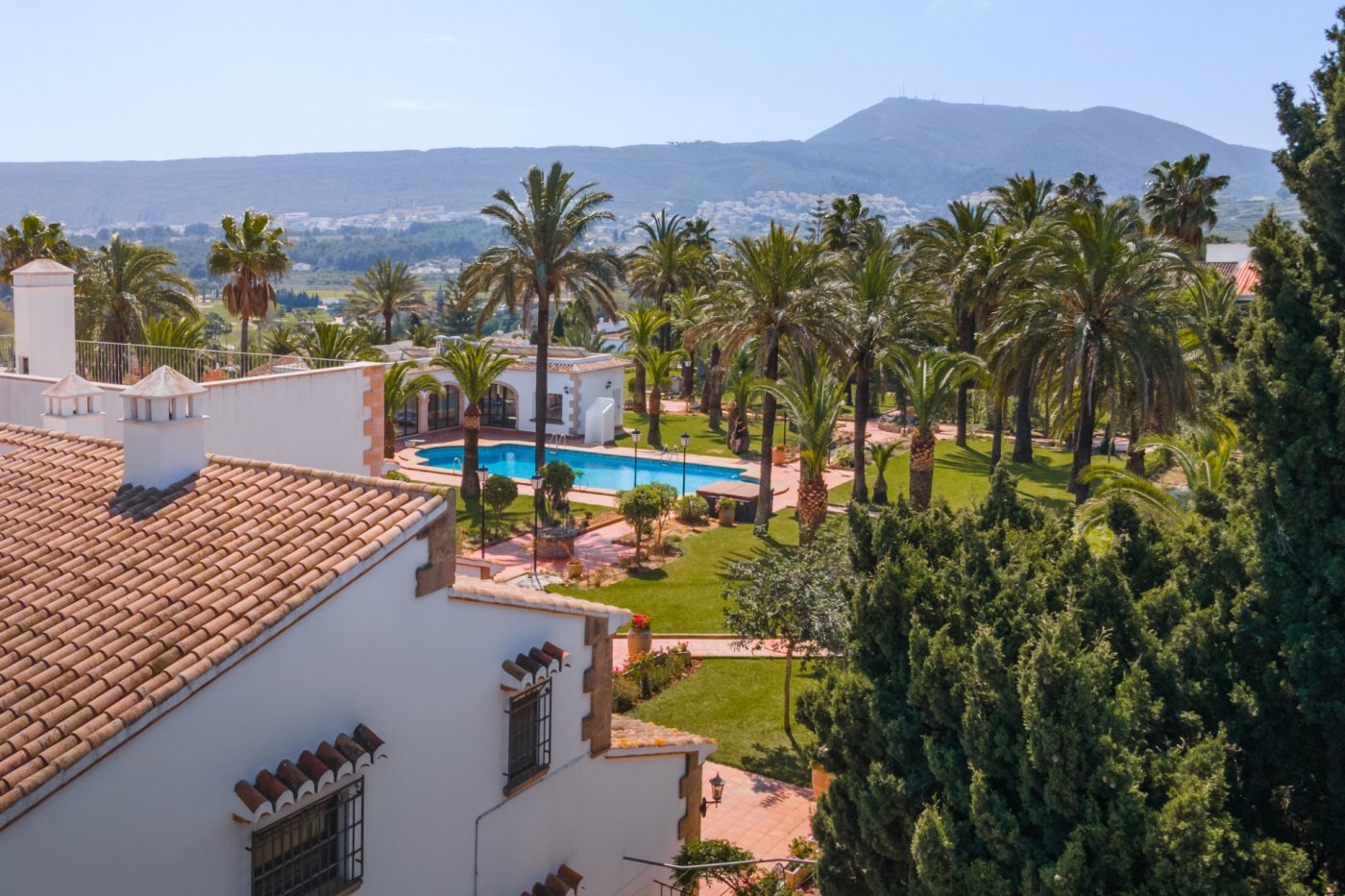 Vista panorámica desde la planta superior que muestra el extenso jardín de palmeras, la piscina de 16x7 metros y la casa de invitados bajo el cielo de Jávea.