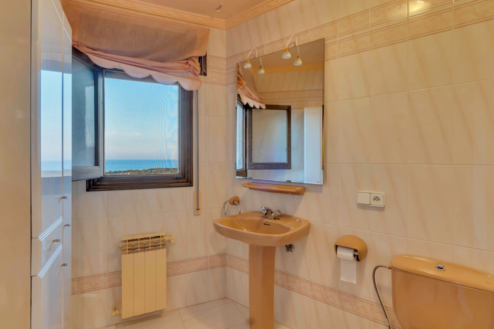 Baño con sanitarios en tono beige, lavabo de pedestal y espejo con luminarias. La ventana ofrece vistas despejadas hacia el mar Mediterráneo.