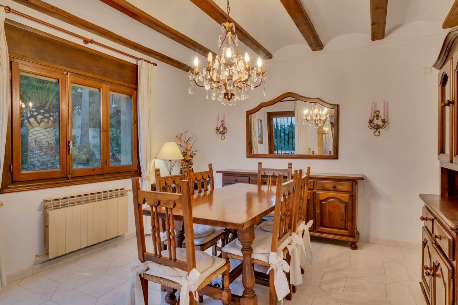 Comedor de estilo rústico con vigas de madera vistas, mobiliario de madera maciza, lámpara de araña de cristal y suelos cerámicos claros.