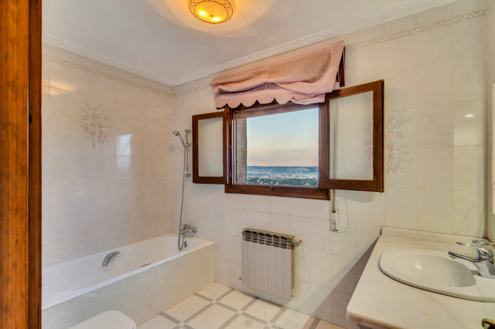 Cuarto de baño con bañera, alicatado con motivos florales y ventana de madera que ofrece vistas despejadas a la bahía y el mar.