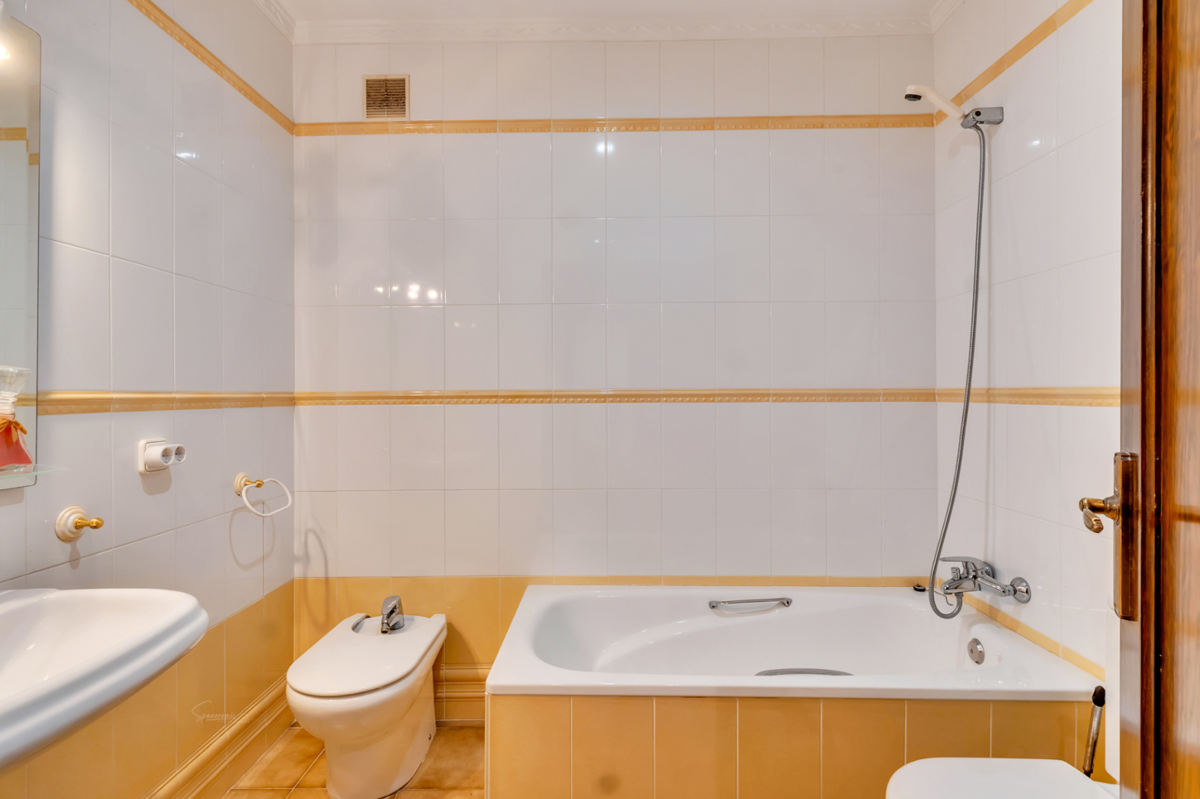 Cuarto de baño funcional con bañera integrada, bidé y revestimiento de azulejos blancos con detalles decorativos en tonos ocre y mostaza.
