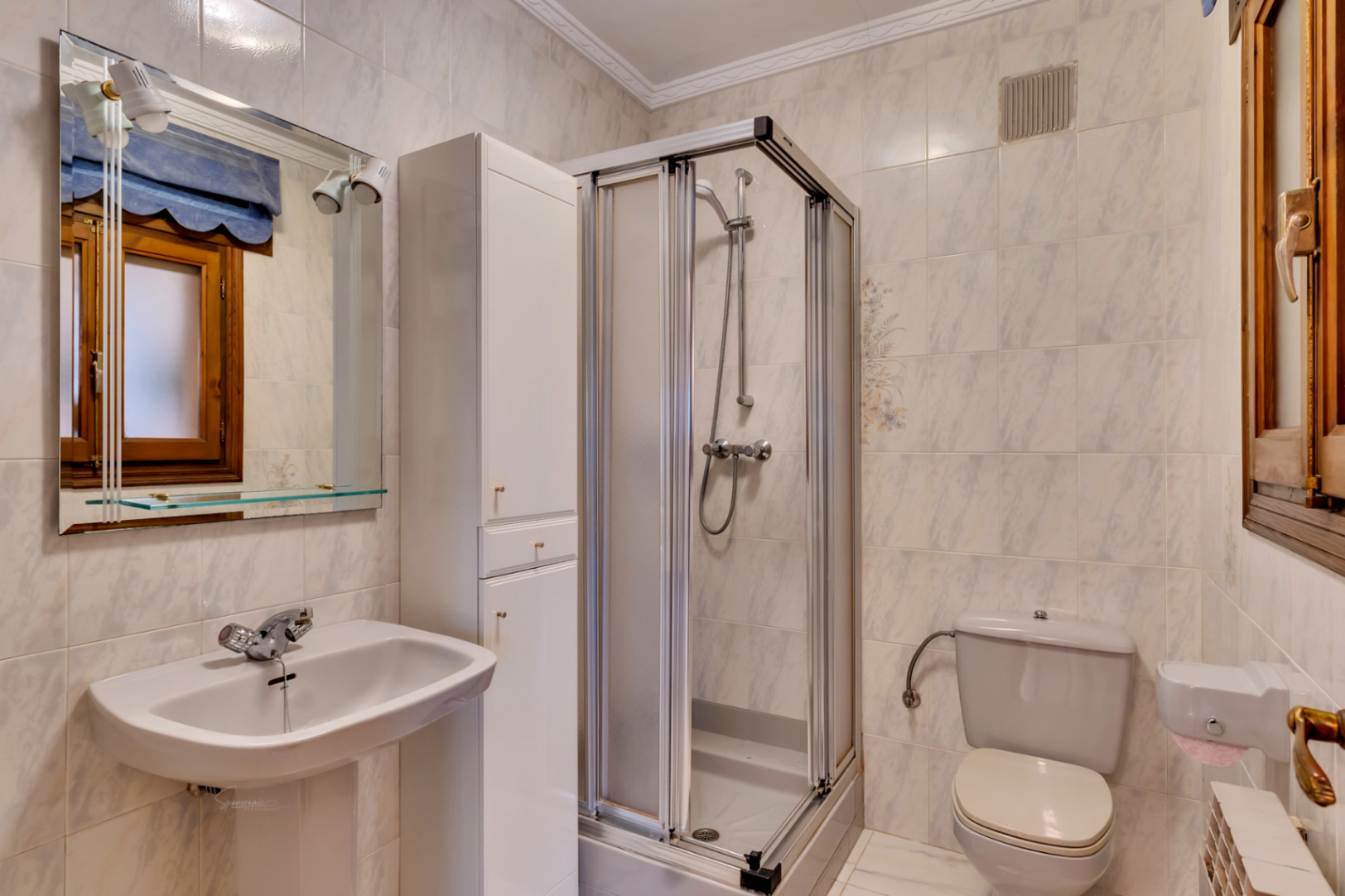Cuarto de baño funcional con lavabo de pedestal, cabina de ducha con puertas correderas y armario auxiliar alto. Paredes alicatadas con azulejos de efecto mármol y ventana de madera.