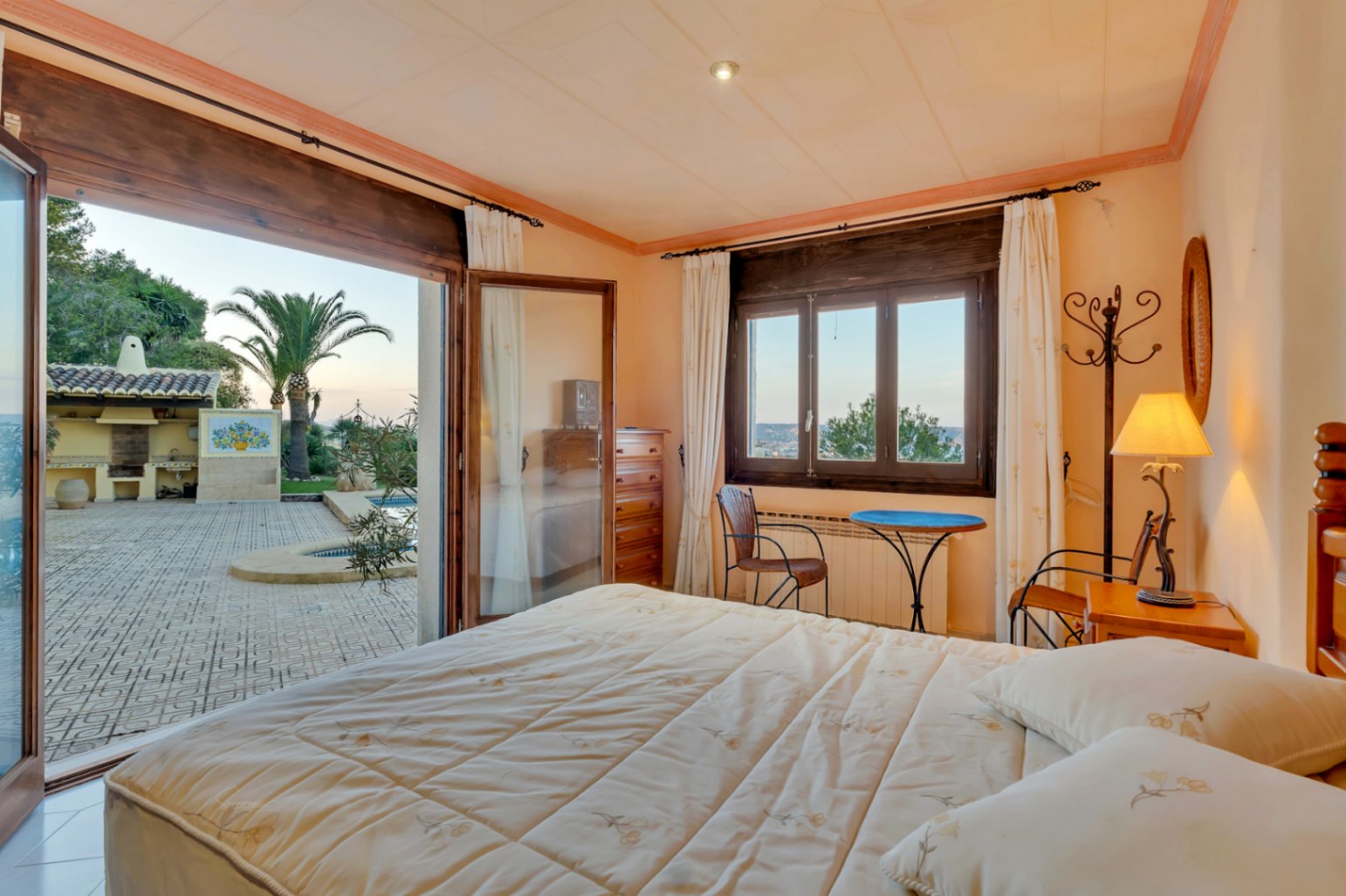 Dormitorio con acceso directo a la terraza, marcos de madera oscura y vistas al jardín mediterráneo con piscina y zona de barbacoa exterior.
