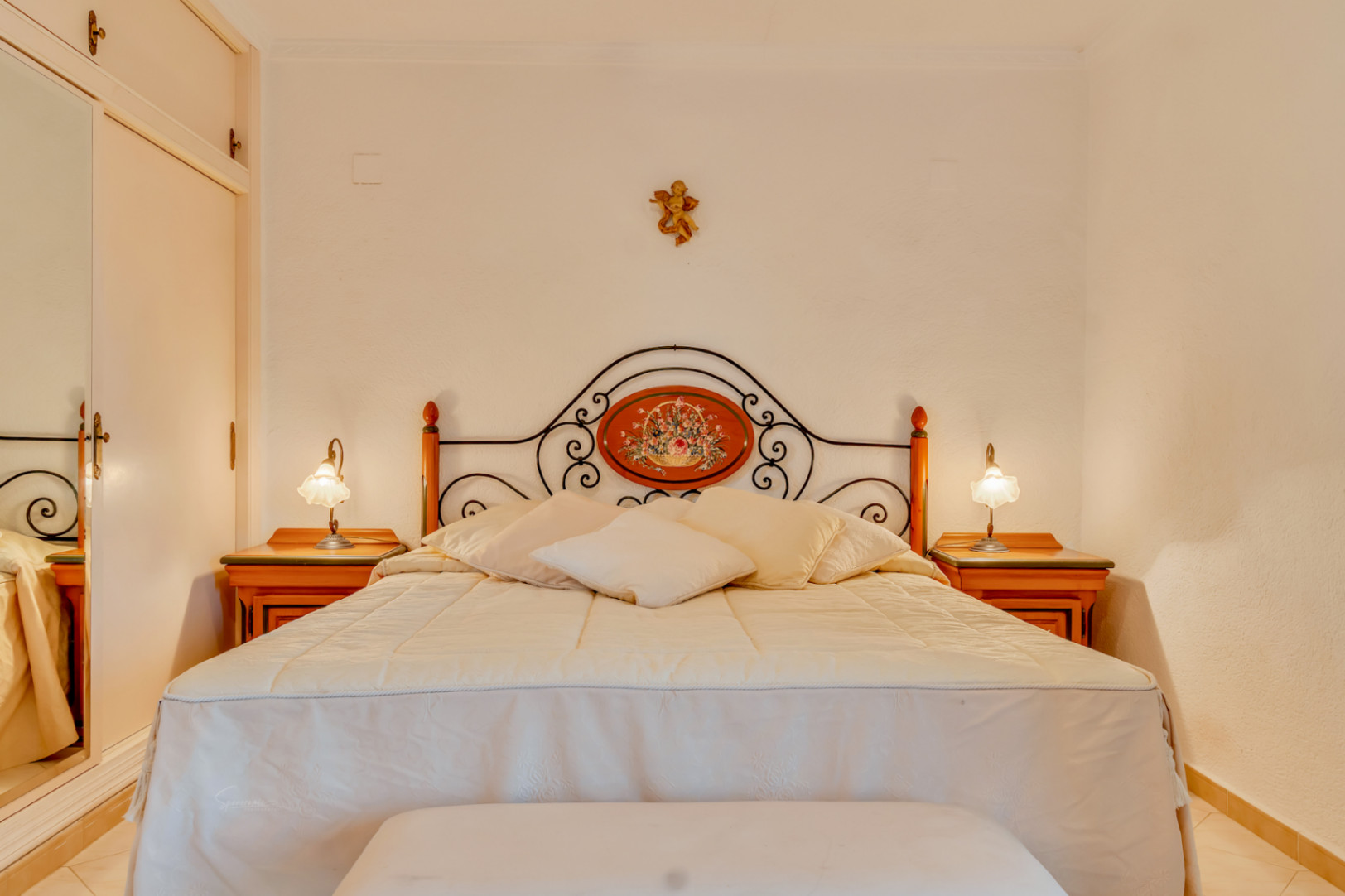Dormitorio con cama doble, cabecero de forja con medallón floral y mesitas de noche de madera. Incluye armario empotrado con espejo lateral.