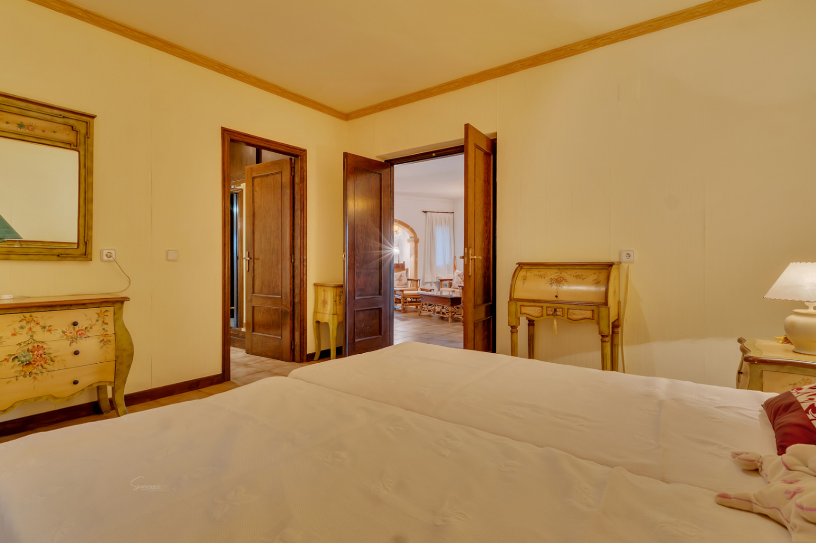 Dormitorio con mobiliario de estilo clásico pintado a mano, puertas dobles de madera oscura y acceso directo a la zona de estar principal.