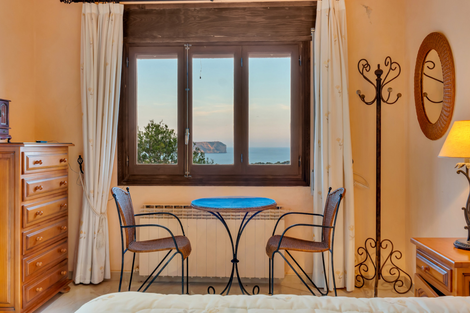 Dormitorio con zona de estar junto a una ventana de madera que ofrece vistas al Cabo San Antonio. Incluye radiador, cómoda de madera y espejo de mimbre.