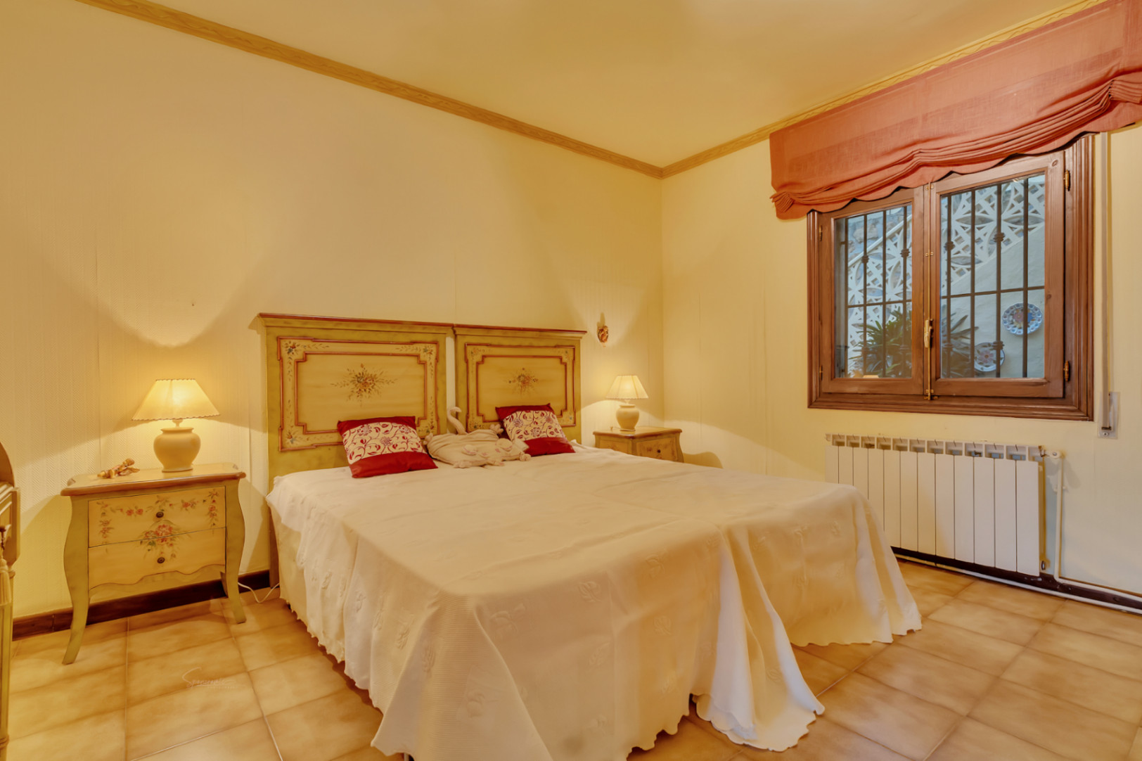 Dormitorio doble con cabeceros de madera pintados a mano, mesitas de noche a juego, suelos de terracota y ventana con marco de madera y calefacción central.