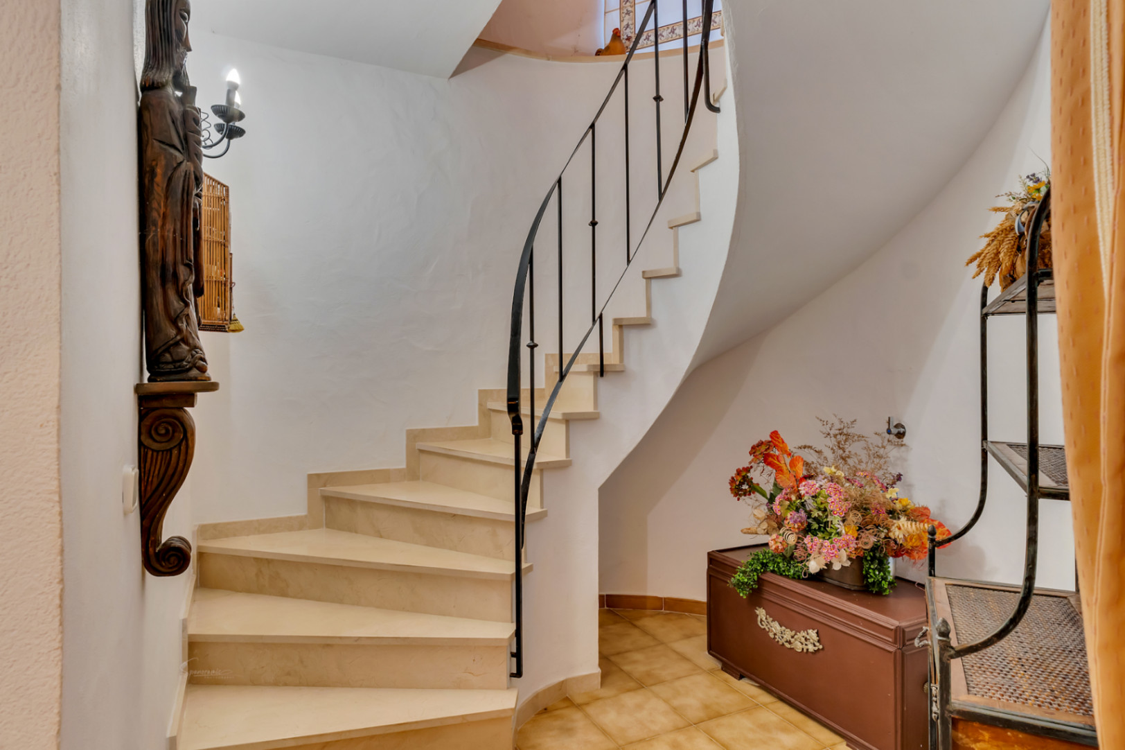 Escalera interior curva con peldaños de piedra clara y barandilla de hierro forjado. El espacio incluye suelos cerámicos y elementos decorativos de estilo rústico.