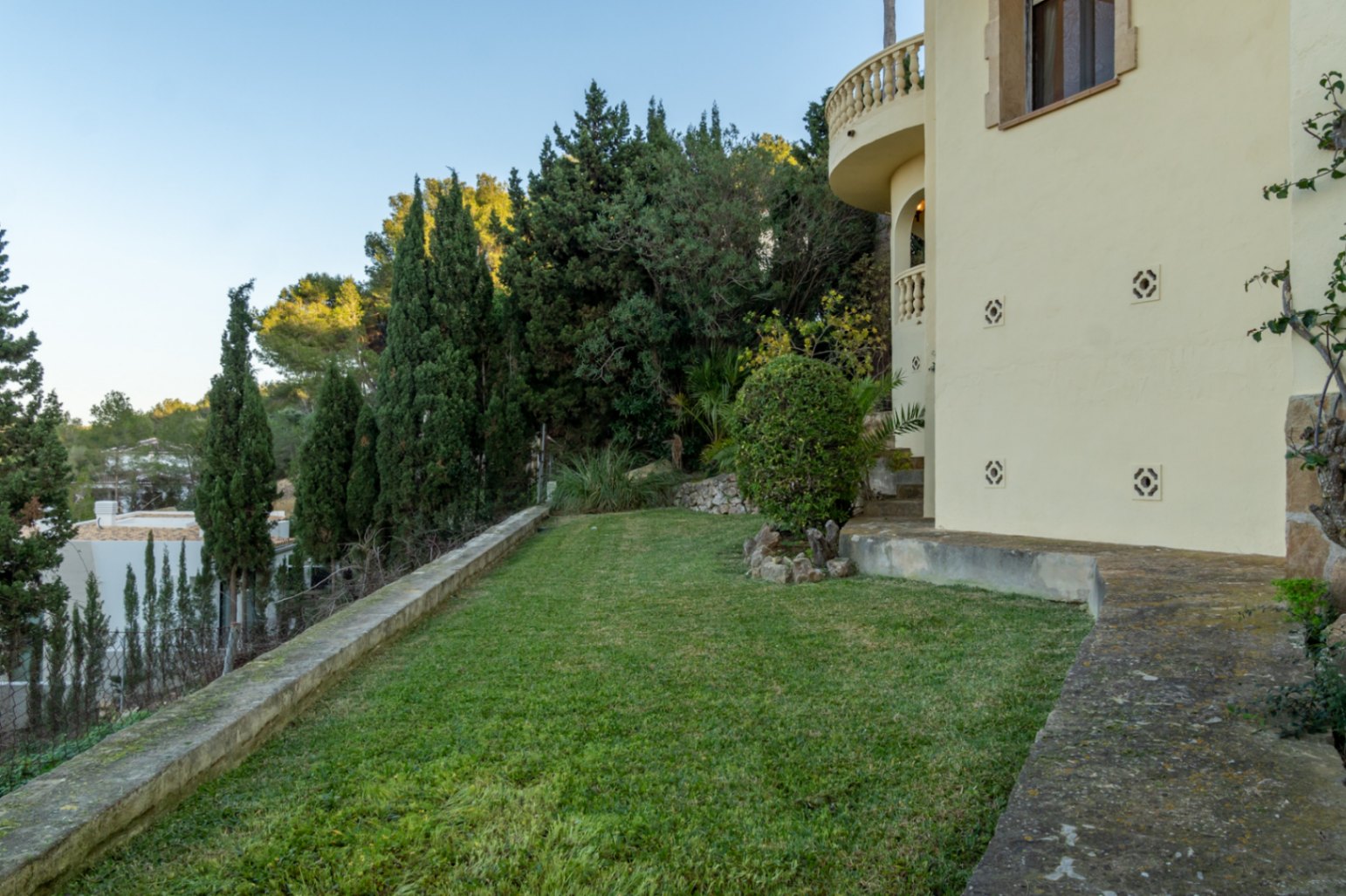 Jardín lateral con césped cuidado, rodeado de cipreses y vegetación autóctona junto a la fachada de la villa con balcones de balaustrada.