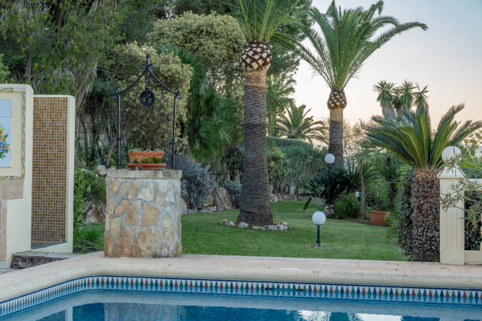 Jardín mediterráneo con césped y palmeras junto a la piscina. Incluye un pozo decorativo de piedra y una ducha exterior revestida con azulejos de mosaico.