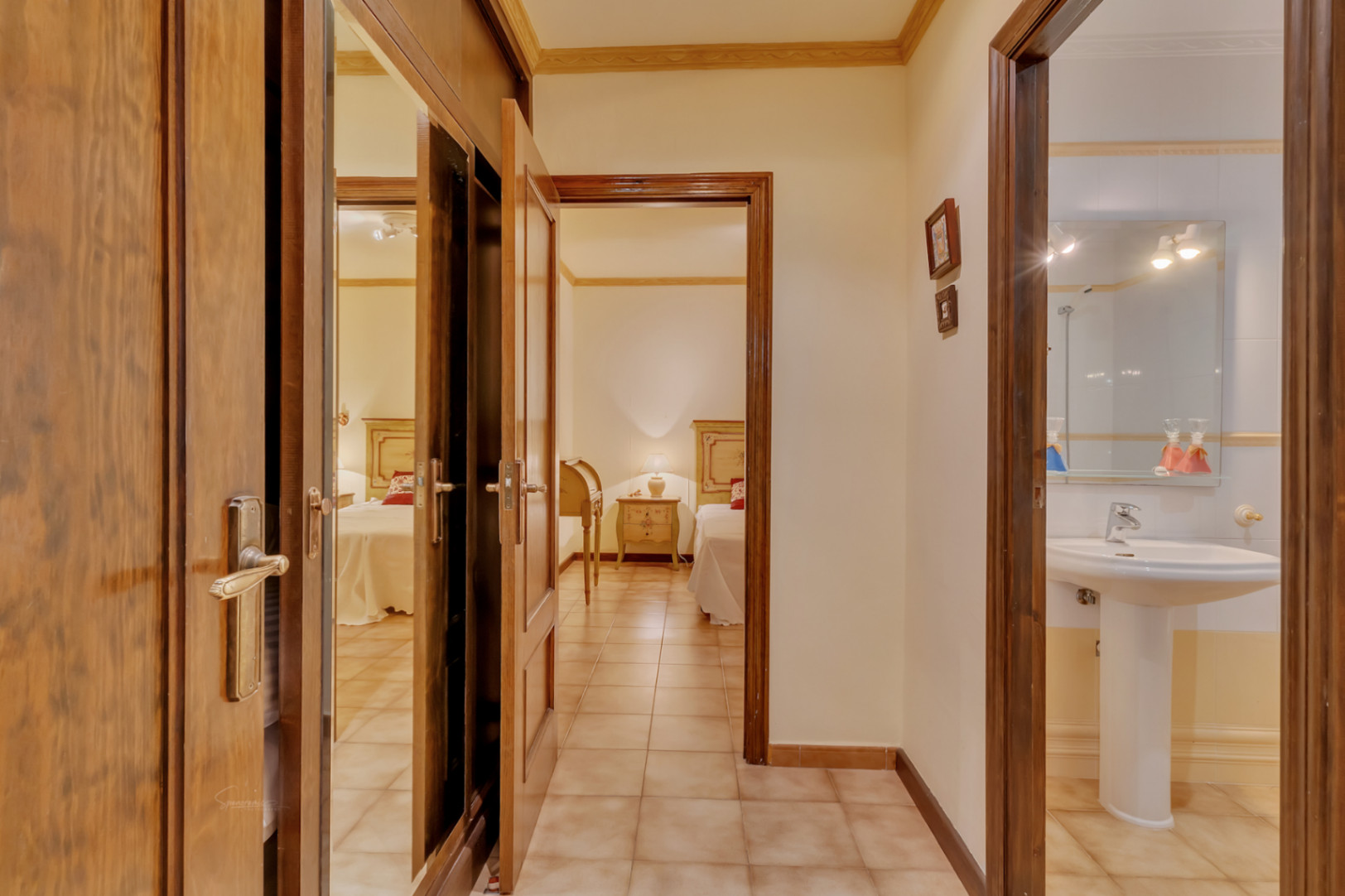 Pasillo distribuidor con suelos cerámicos que conecta un dormitorio con camas individuales, un baño con lavabo de pedestal y armarios empotrados de madera con espejos.