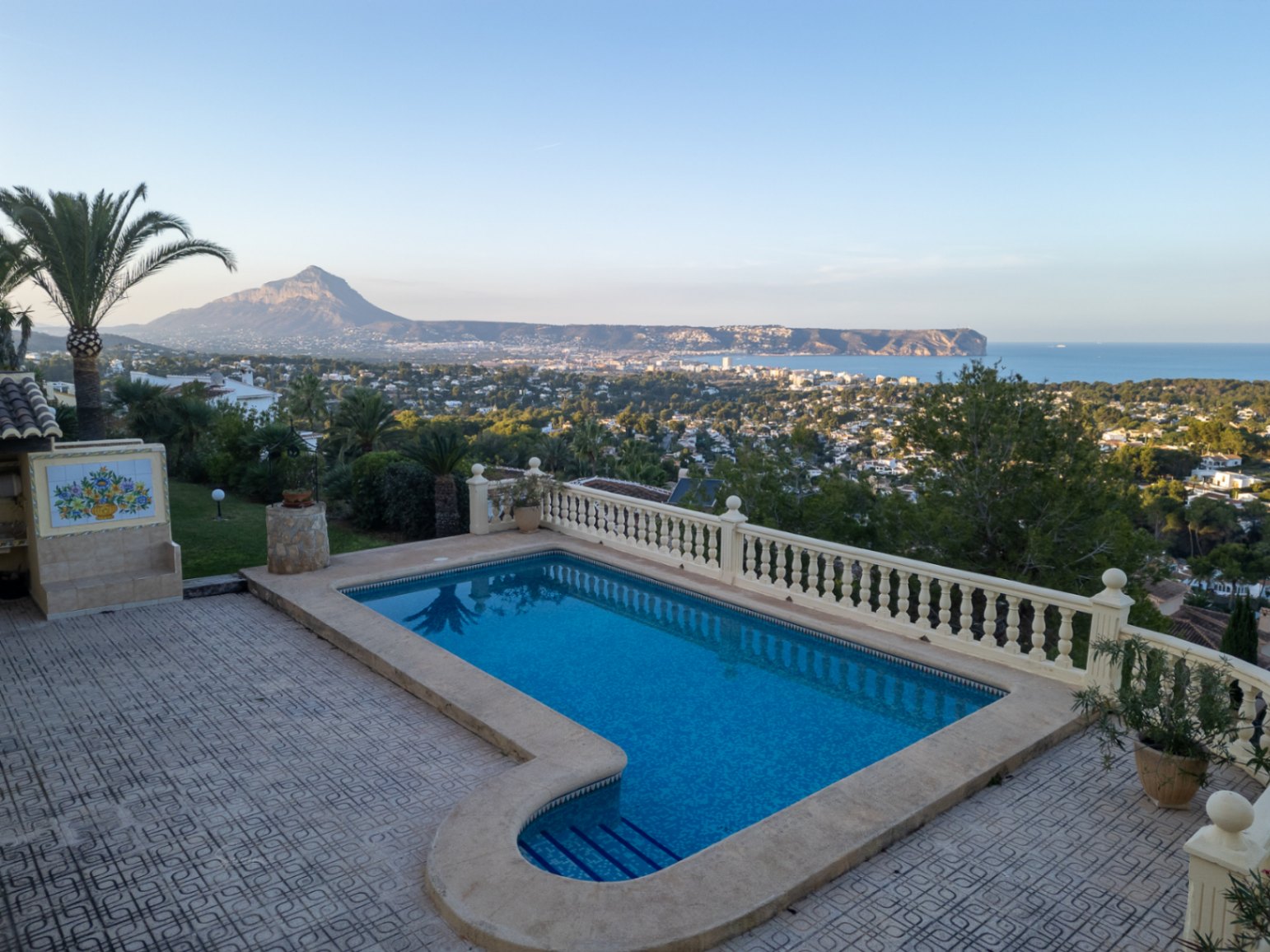 Piscina de 8x4 metros con vistas panorámicas a la bahía de Jávea y el Montgó, rodeada de una amplia terraza con balaustrada clásica y jardín mediterráneo.