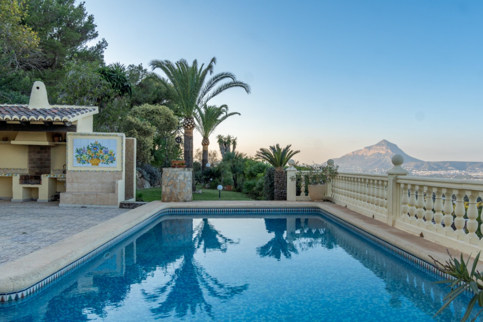 Piscina de mosaico azul junto a una zona de barbacoa techada con mural cerámico, rodeada de jardines mediterráneos y vistas al Montgó.