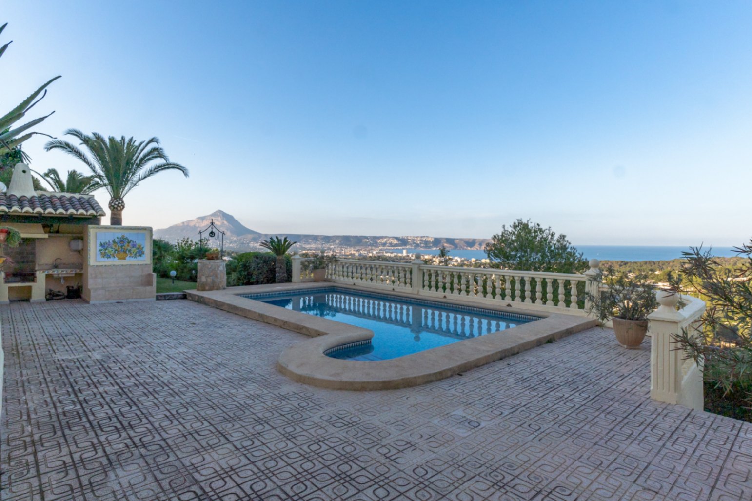 Piscina exterior con terraza de gres y balaustrada, ofreciendo vistas panorámicas al Montgó y a la bahía de Jávea. Incluye zona de barbacoa y jardín mediterráneo.
