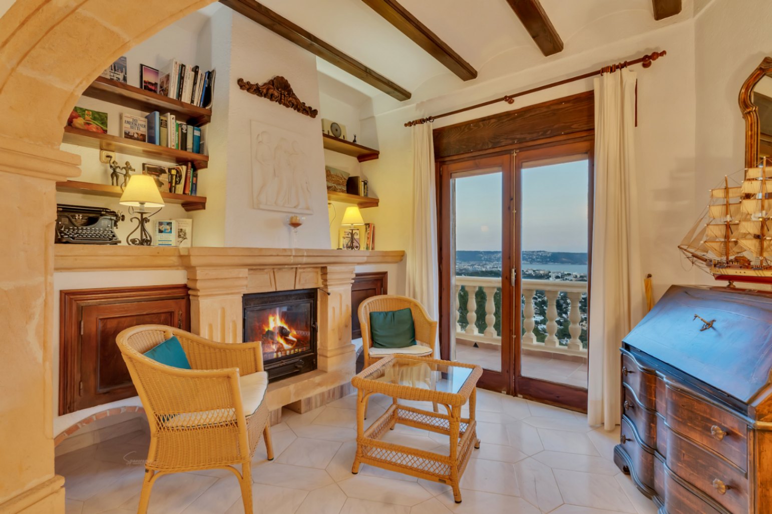 Salón con chimenea de piedra tosca, vigas de madera y escritorio clásico. La puerta acristalada ofrece vistas a la bahía de Jávea y el Cabo San Antonio.