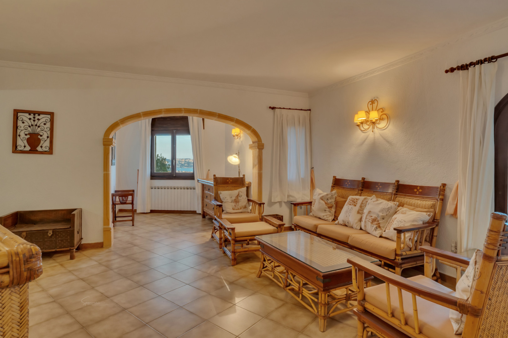 Salón con mobiliario de bambú, suelos cerámicos y un arco de piedra tosca que comunica con una zona de estar con vistas al mar y radiadores.