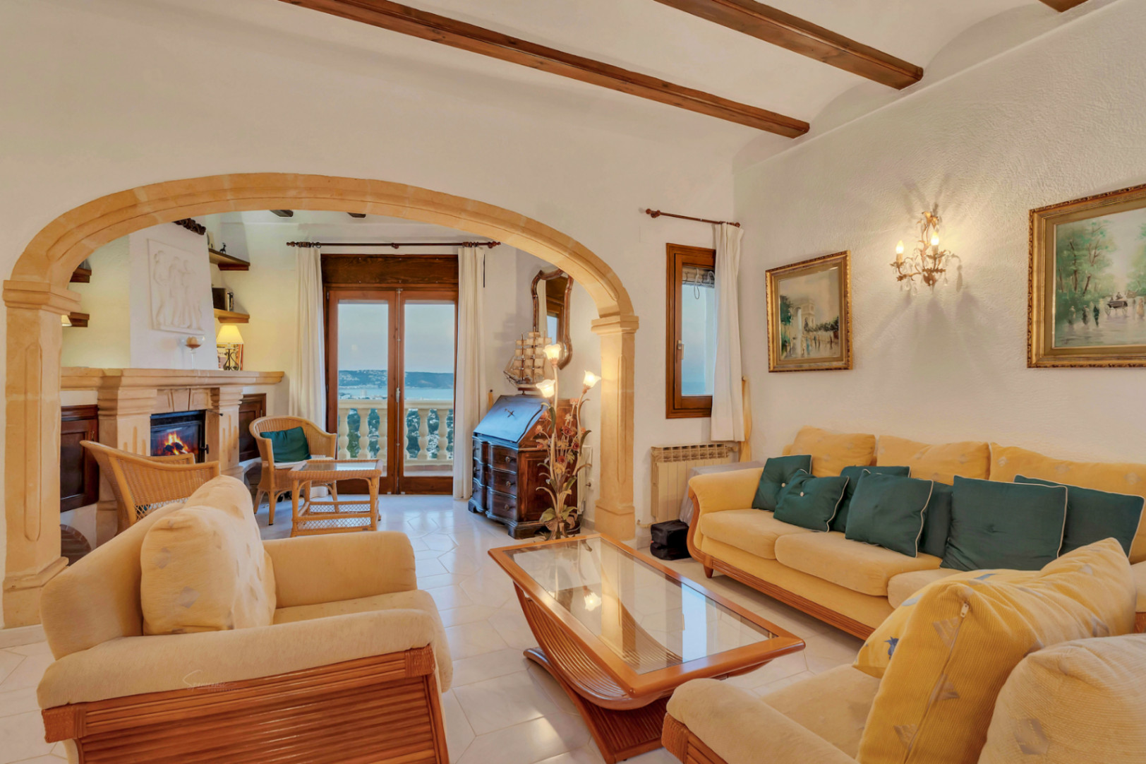 Salón principal con vigas de madera, arco de piedra tosca y chimenea. El espacio cuenta con mobiliario mediterráneo y acceso a la terraza con vistas al mar.