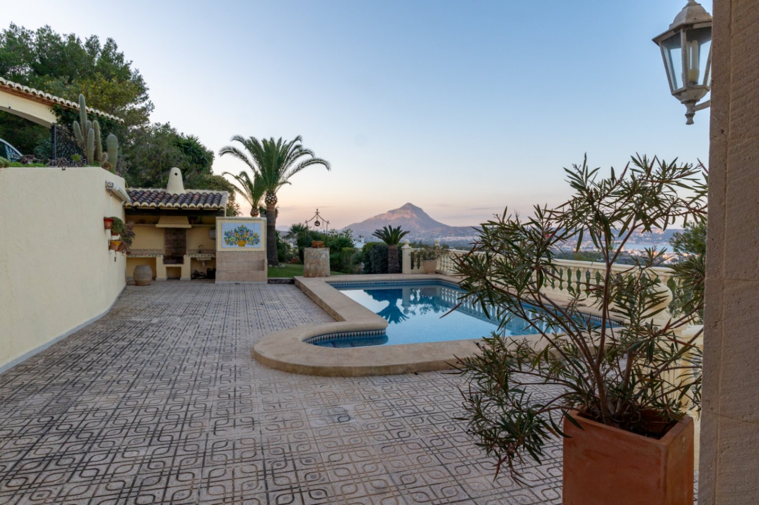 Terraza con piscina, zona de barbacoa de obra y vistas panorámicas al Montgó y la bahía de Jávea. El suelo presenta baldosas con motivos geométricos.