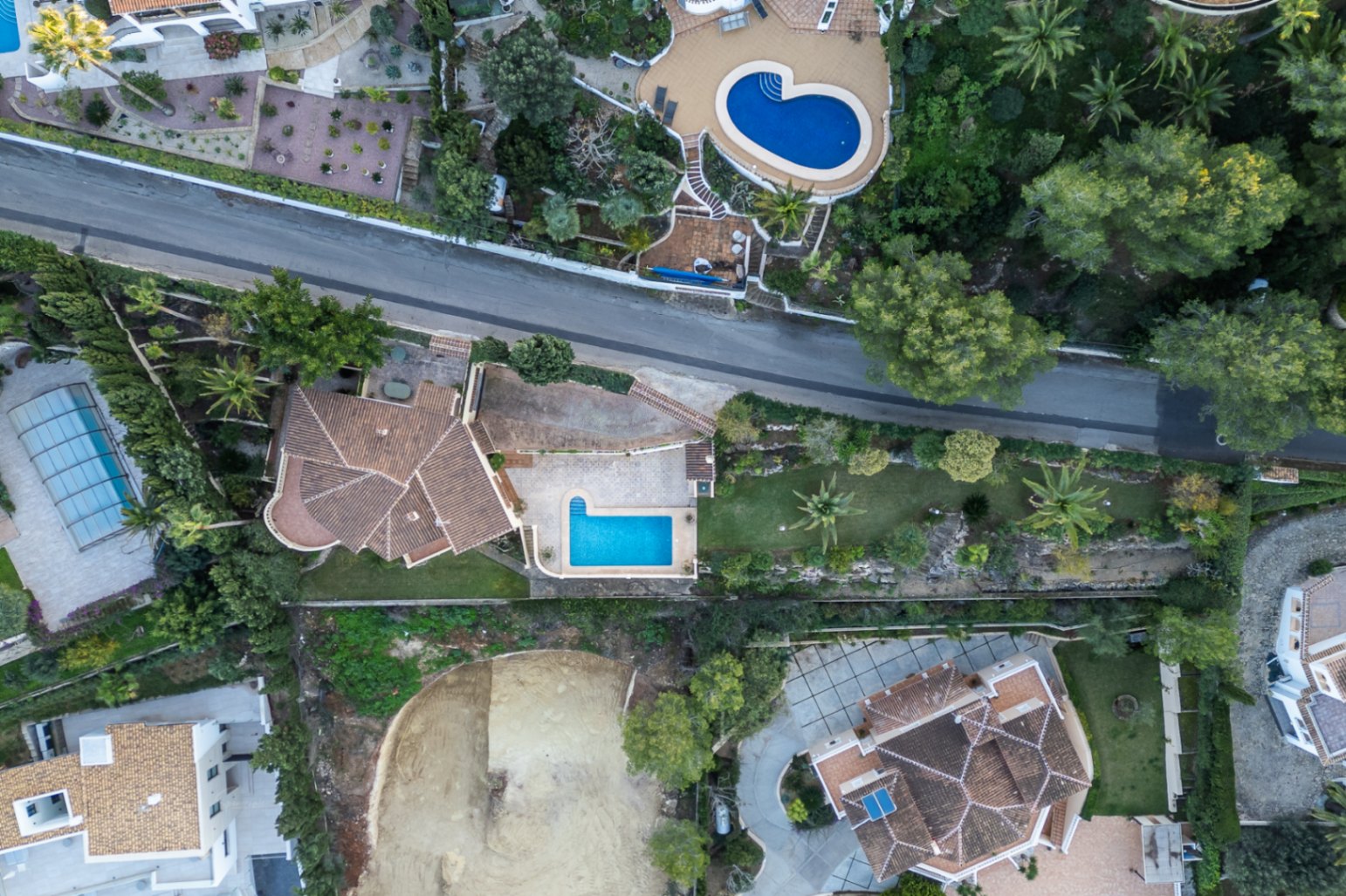 Vista aérea cenital de la propiedad que muestra la distribución de la parcela, la piscina en forma de L, las terrazas pavimentadas y el jardín mediterráneo con palmeras.