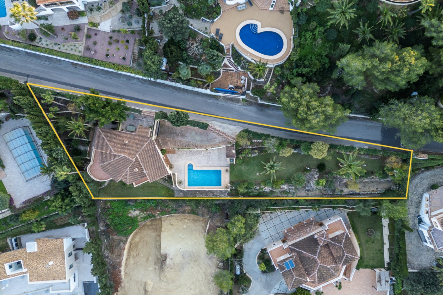 Vista aérea perimetrada de la parcela, destacando la piscina en forma de L, el jardín mediterráneo con palmeras y la arquitectura de la villa con tejados de teja.