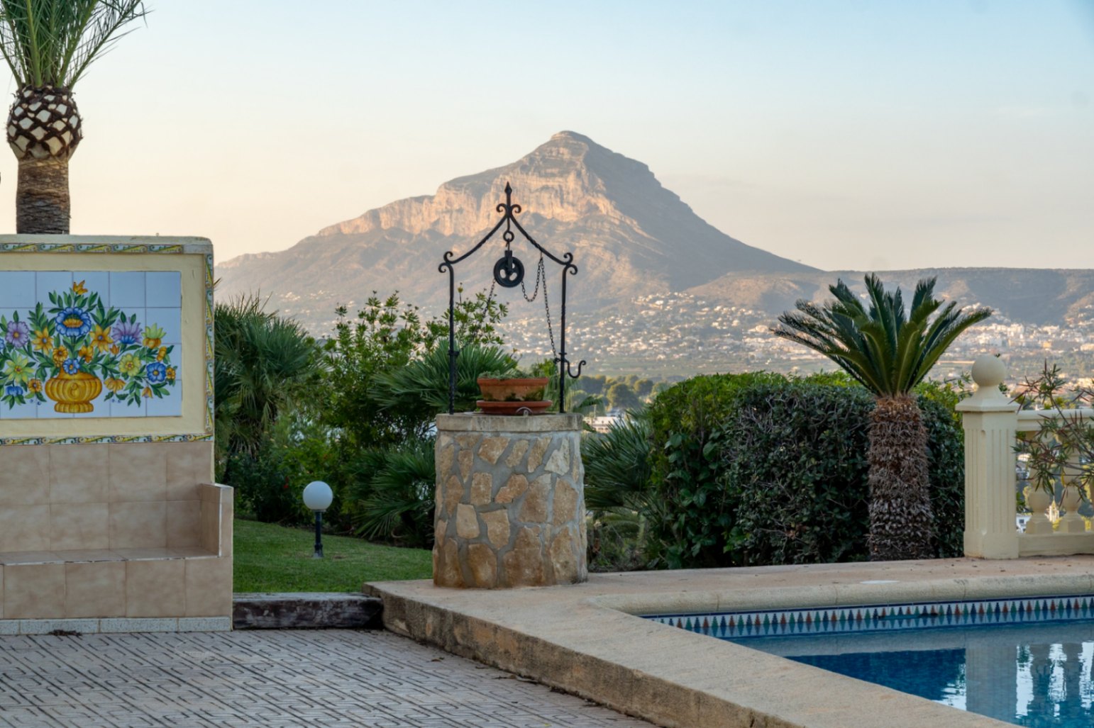Vista desde la piscina hacia el Cabo San Antonio, con un pozo decorativo de piedra y un mural cerámico tradicional rodeado de vegetación mediterránea.