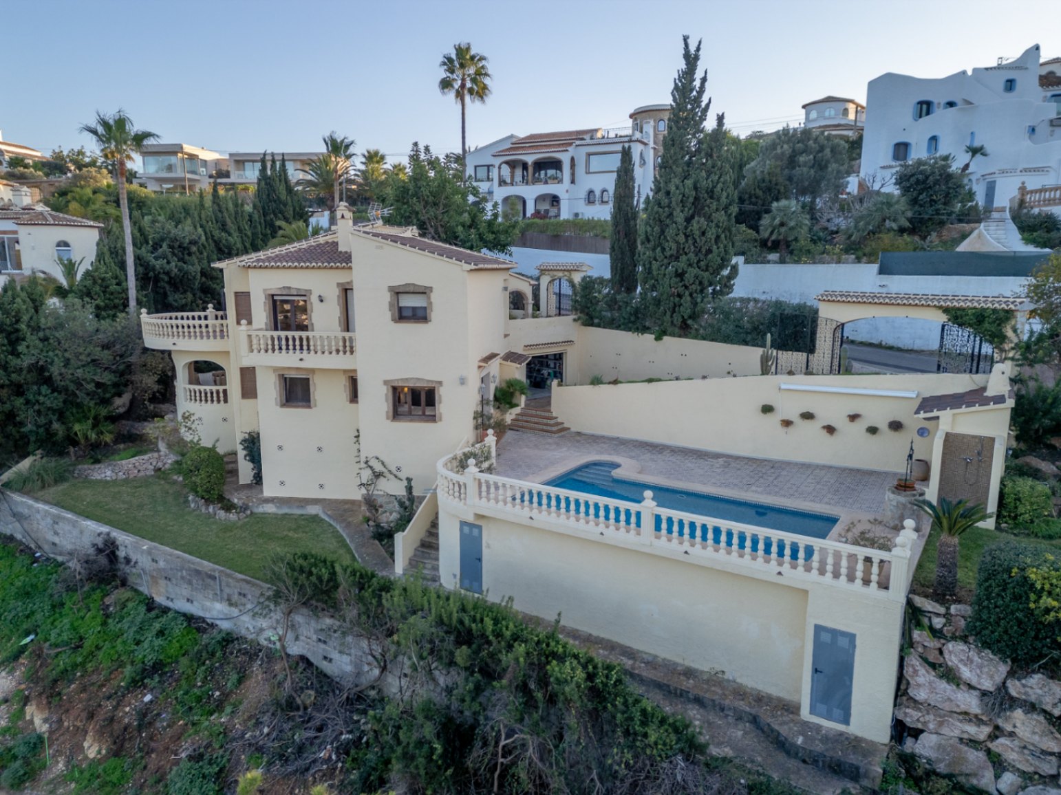 Vista exterior elevada de la villa mediterránea con piscina privada, amplias terrazas con balaustradas blancas y fachada con marcos de piedra natural.