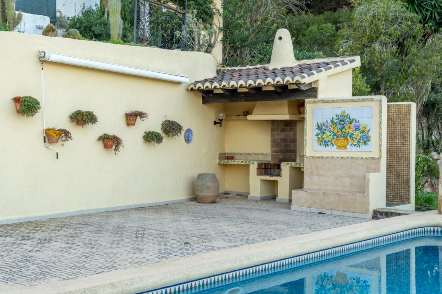 Zona de barbacoa de obra con tejado de teja, mural cerámico decorativo y ducha exterior junto a la piscina con suelo de gres estampado.