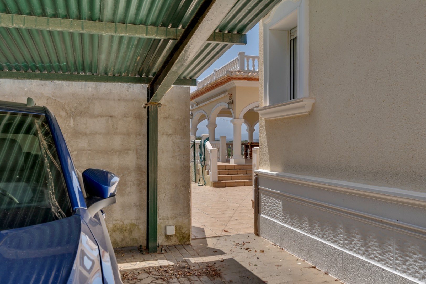 Área de estacionamiento cubierto con marquesina metálica junto a la fachada principal, con acceso directo a la terraza elevada y la naya de arcos tradicionales.