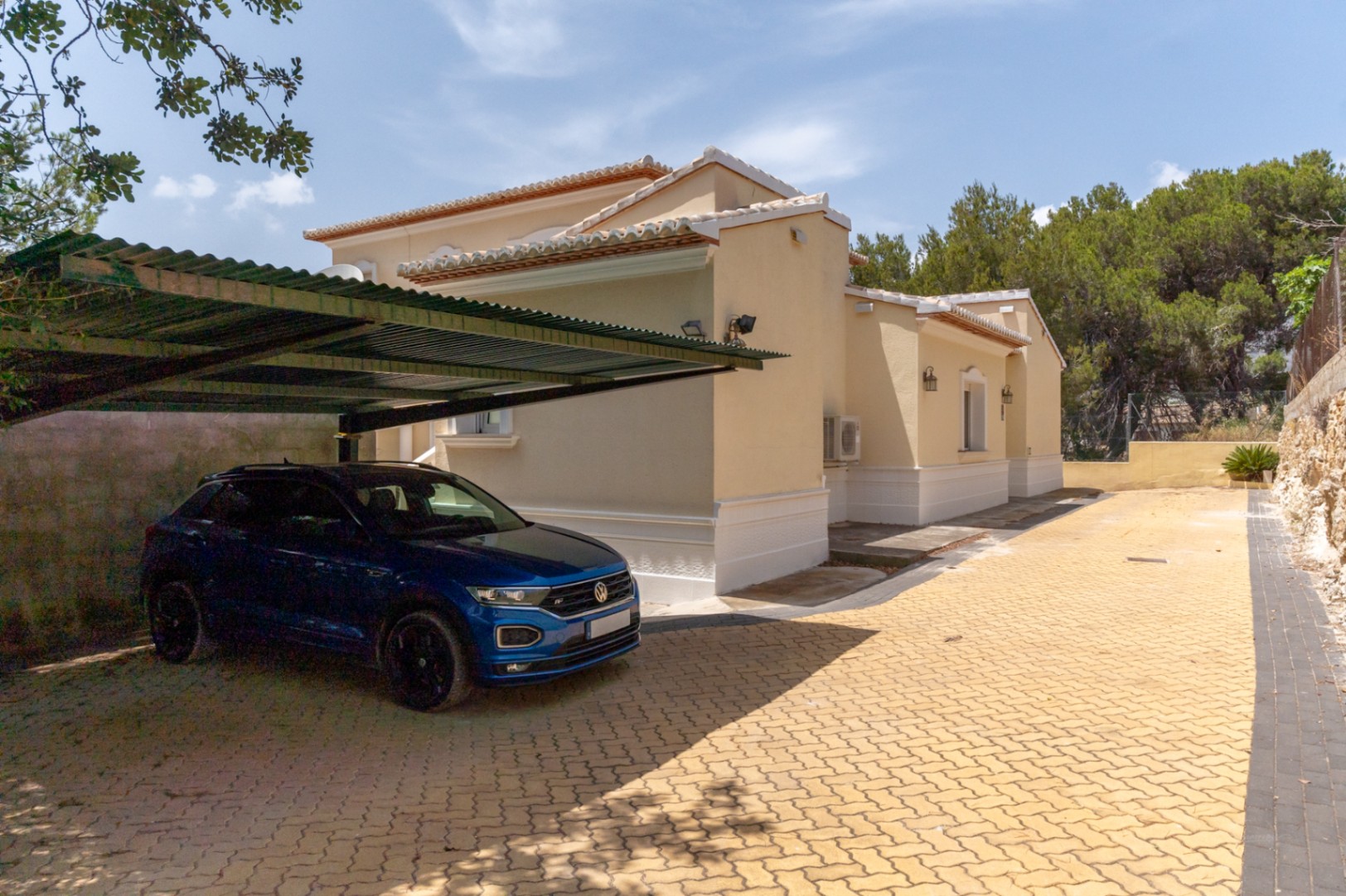 Área de estacionamiento exterior con suelo de adoquines amarillos y una estructura de aparcamiento cubierto junto a la fachada lateral de la villa.