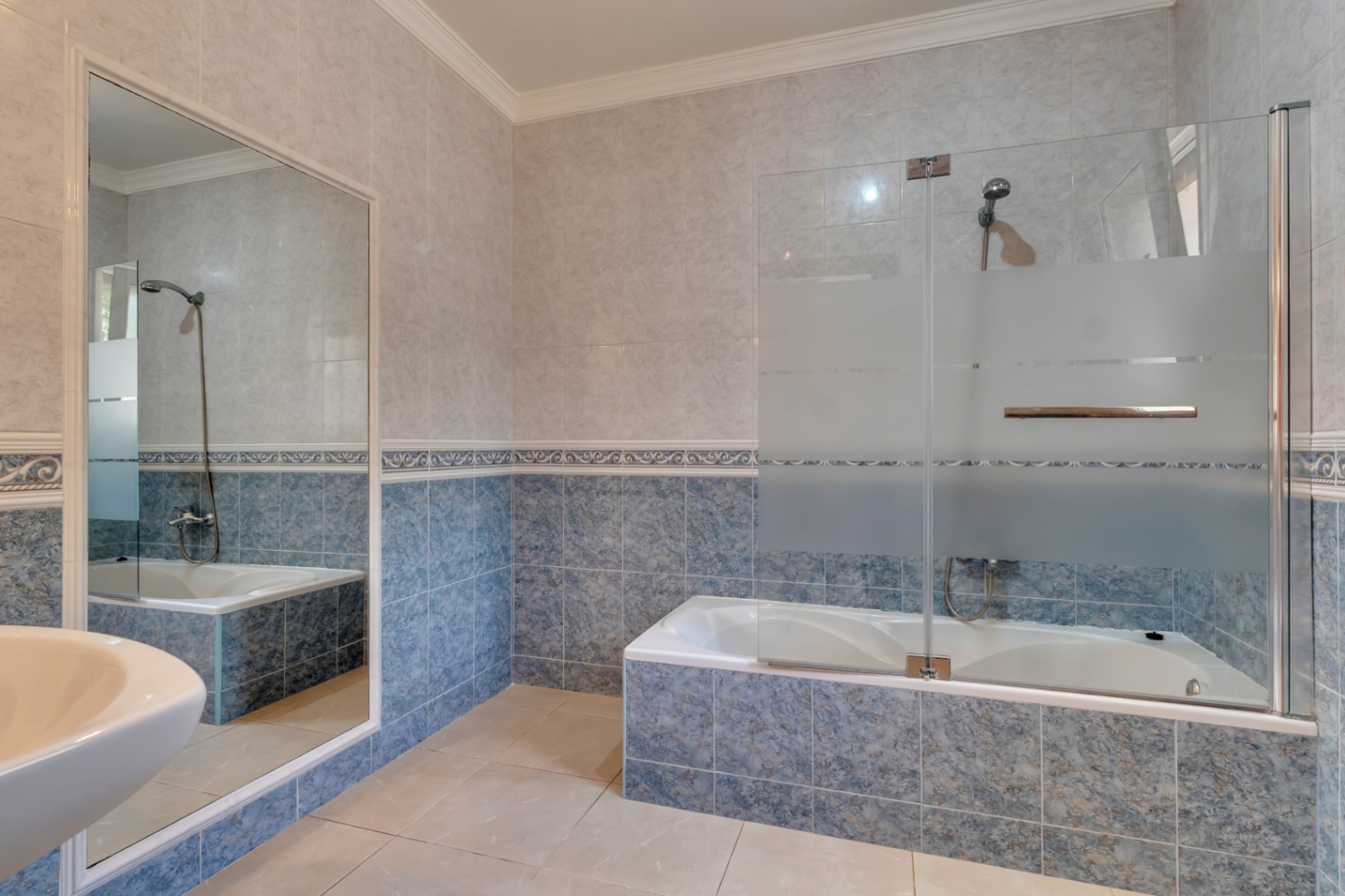 Baño con bañera integrada, mampara de cristal parcialmente esmerilado y revestimiento de azulejos en tonos azules y beige con cenefa decorativa.
