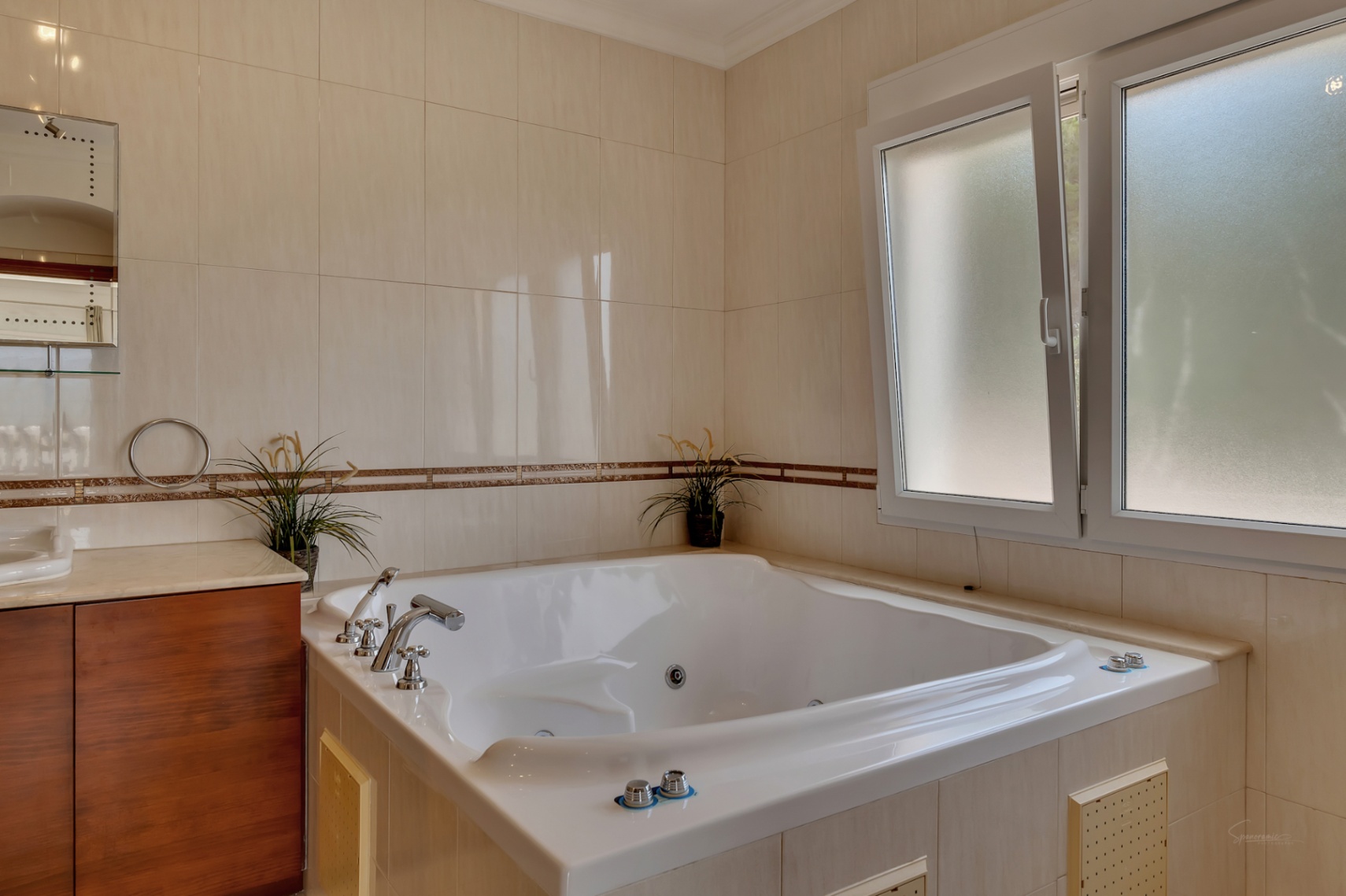Cuarto de baño con bañera de hidromasaje encastrada, paredes alicatadas en tonos crema con cenefa decorativa y ventana de cristal translúcido que aporta luz natural.