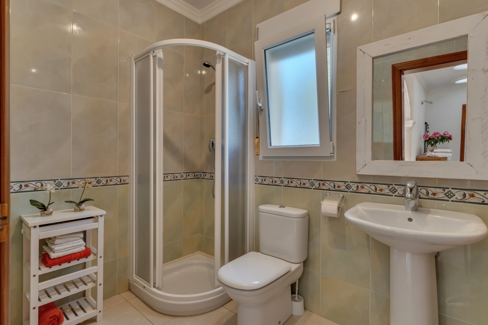 Cuarto de baño con ducha de esquina, lavabo de pedestal e inodoro, con paredes alicatadas en tonos neutros, cenefa decorativa y ventana para luz natural.