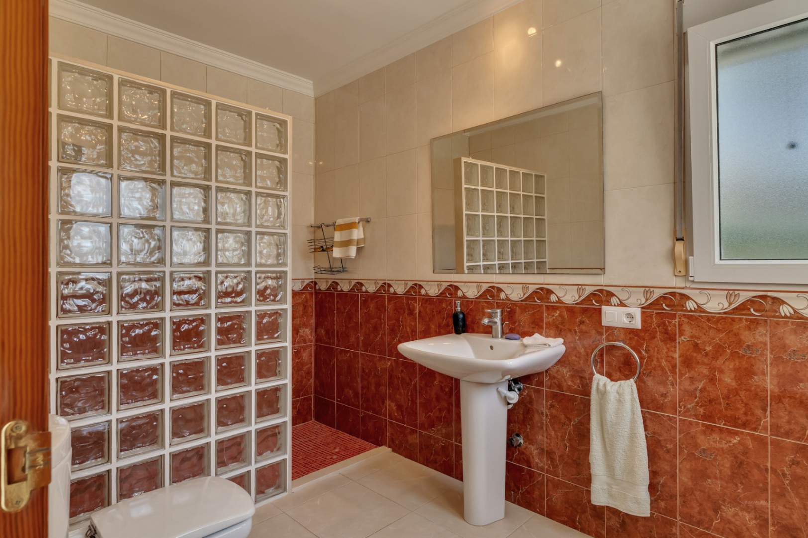 Cuarto de baño con lavabo de pedestal, espejo de pared y una zona de ducha delimitada por una pared de bloques de vidrio y azulejos en tonos rojizos y crema.