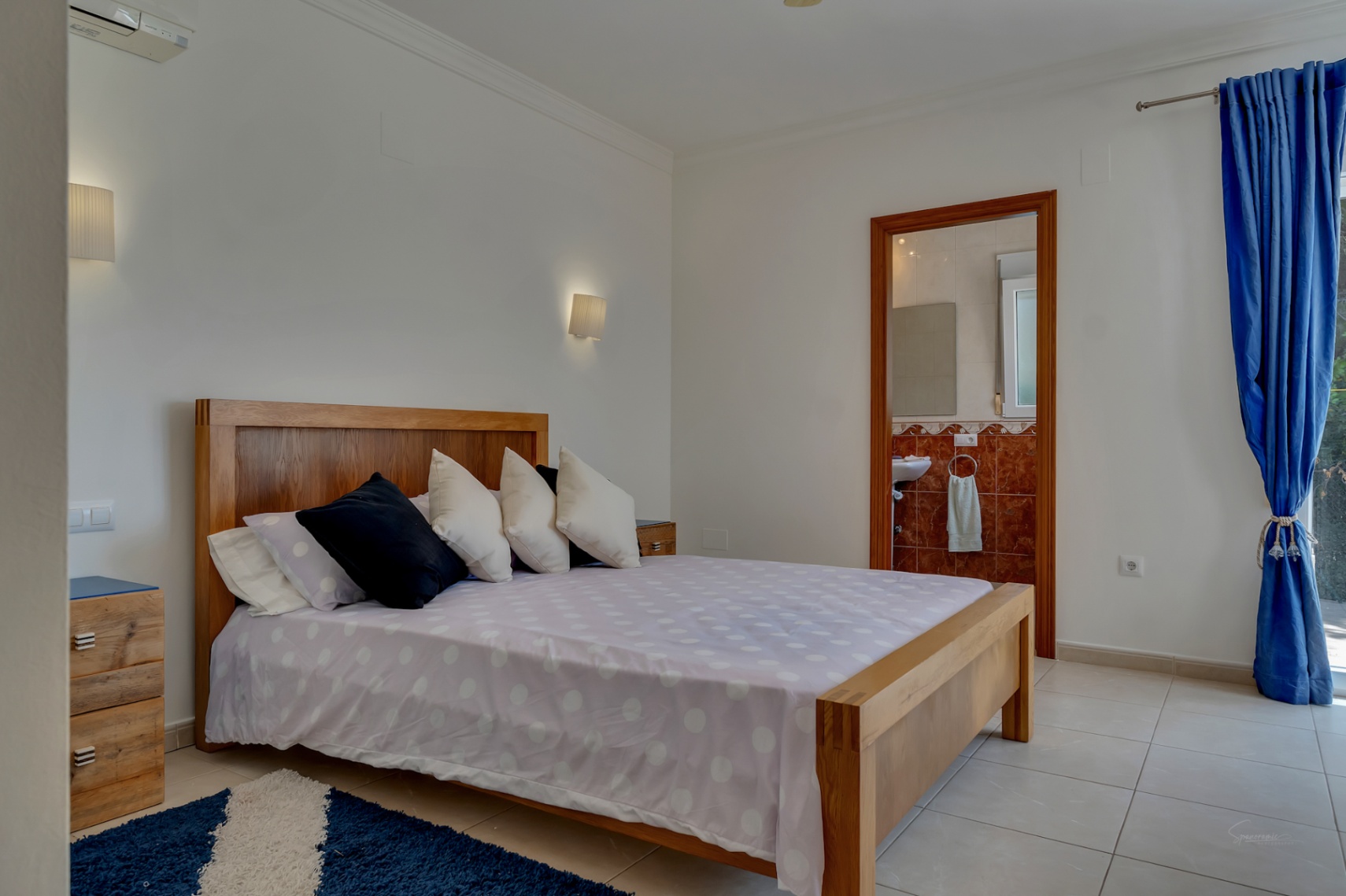 Dormitorio con cama de madera, suelo cerámico y baño en suite con azulejos rojizos. Dispone de aire acondicionado, iluminación de pared y acceso exterior.