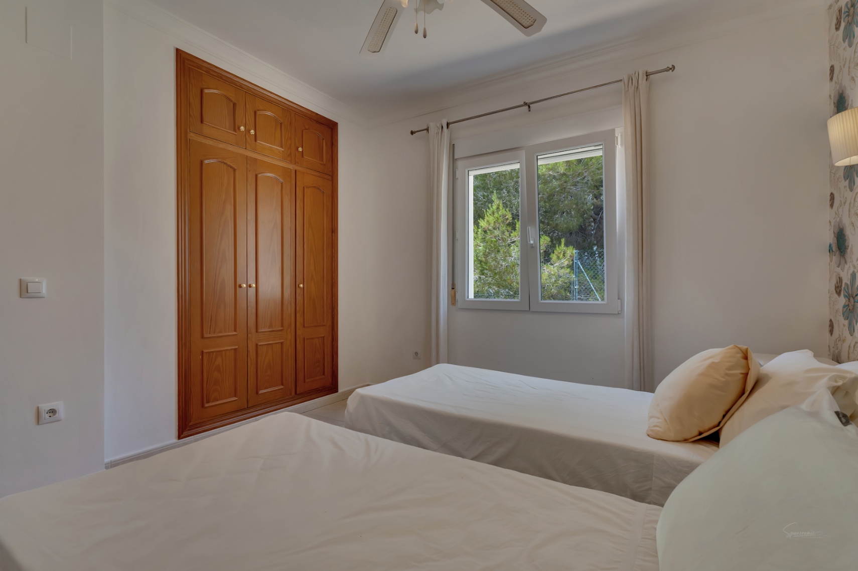 Dormitorio con dos camas individuales, armario empotrado de madera, ventilador de techo y ventana con vistas a la vegetación exterior.