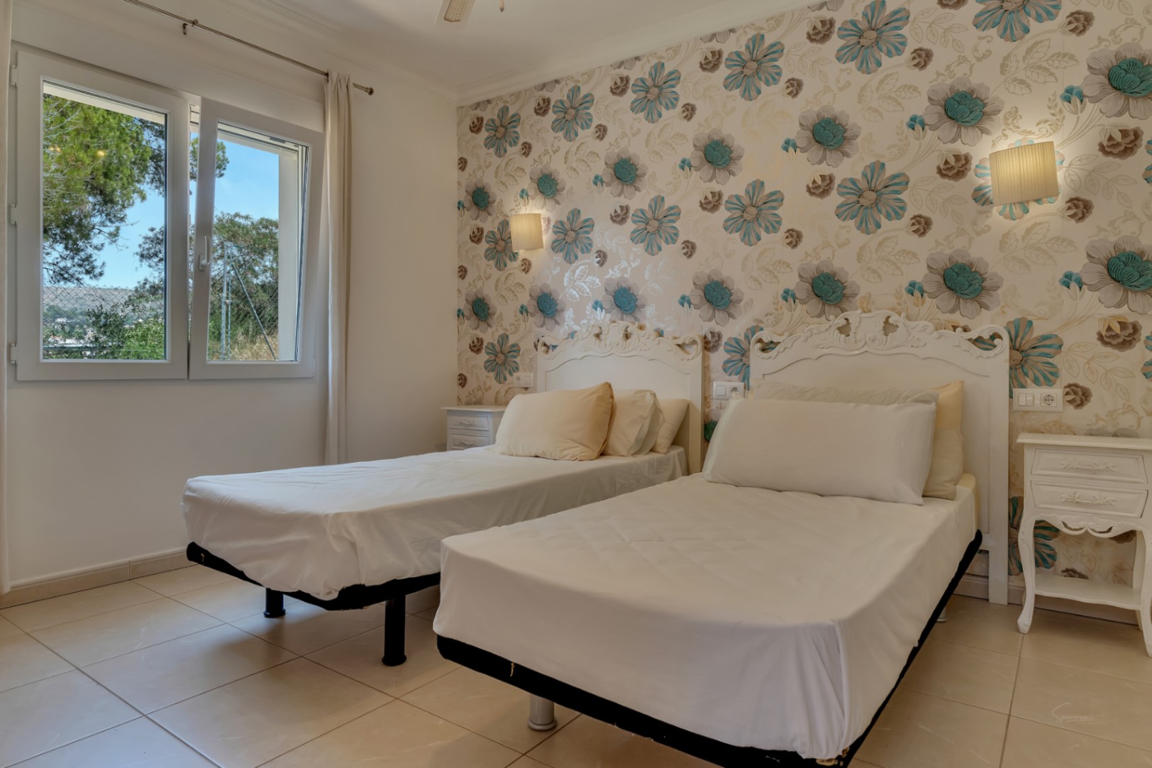 Dormitorio con dos camas individuales, cabeceros blancos ornamentados y papel pintado floral. Cuenta con suelos cerámicos y ventana con vistas a la vegetación exterior.
