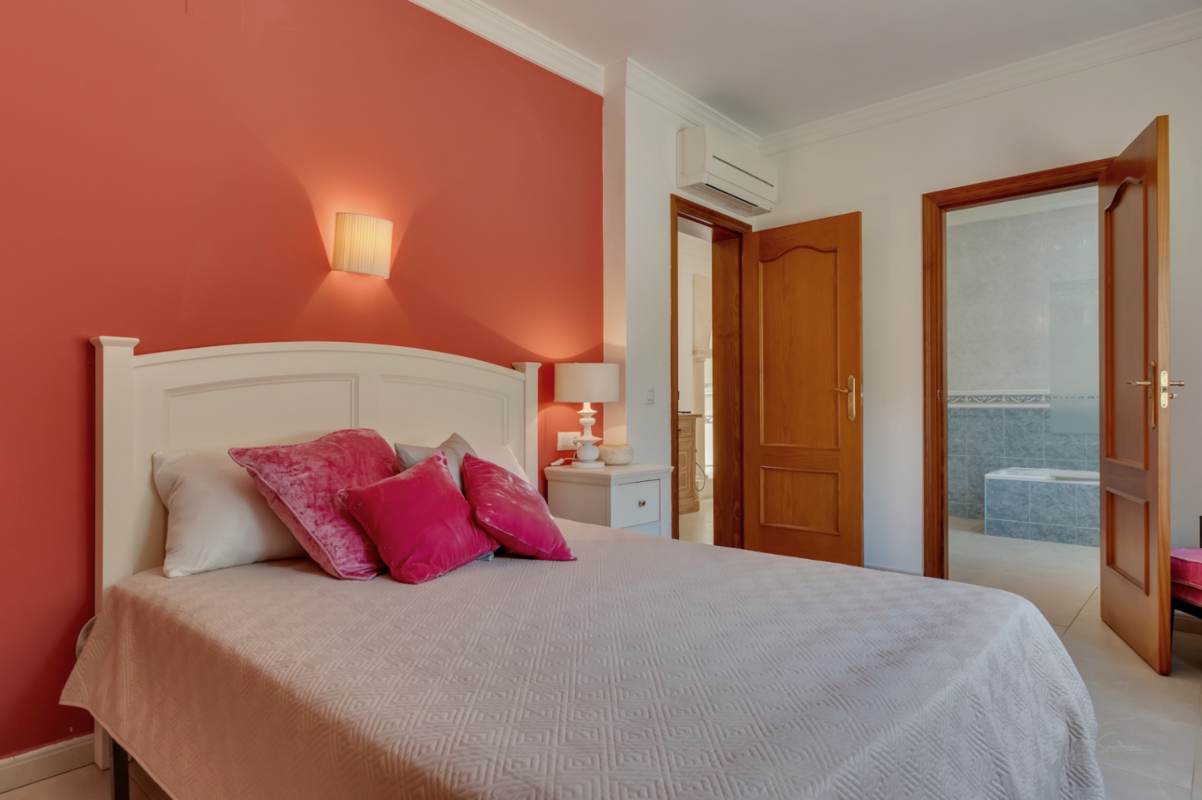 Dormitorio con pared de acento roja, cabecero blanco y aire acondicionado. Incluye baño en suite con azulejos azules y bañera, accesible mediante puertas de madera maciza.