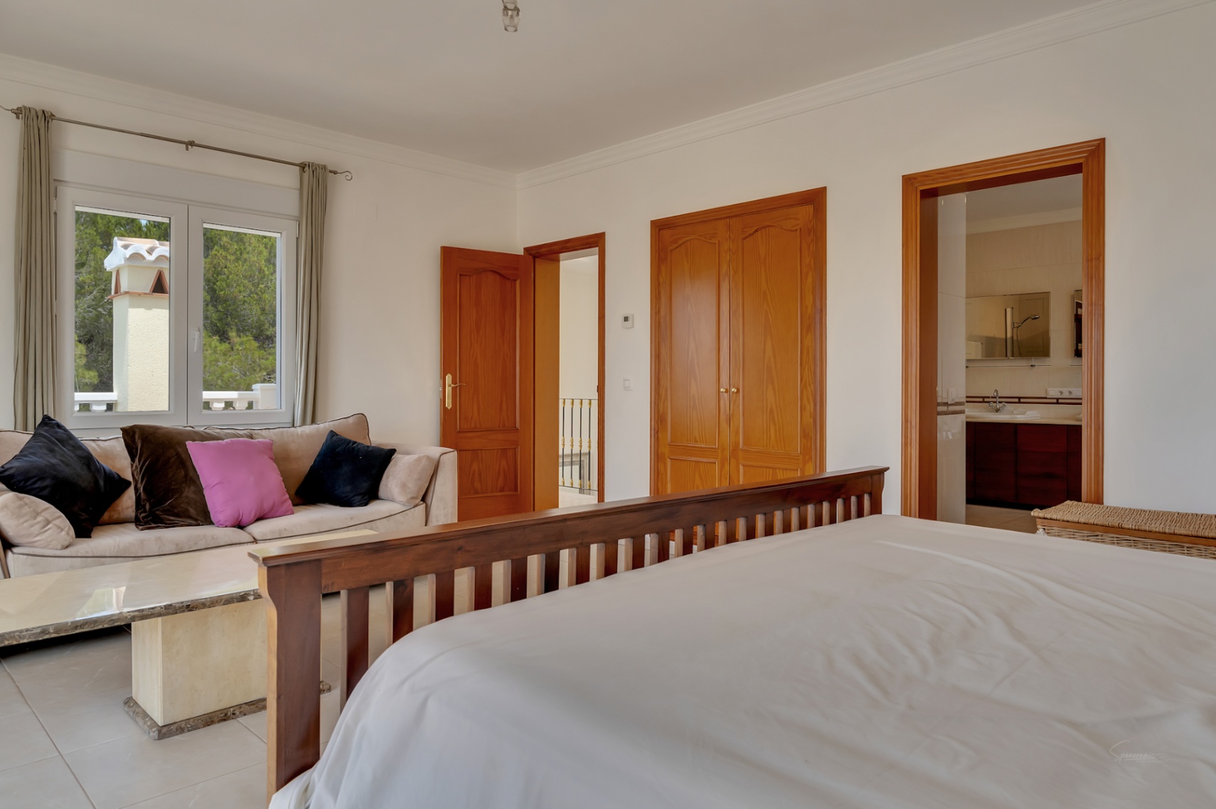 Dormitorio principal con zona de estar, armarios empotrados de madera y baño en suite. La estancia cuenta con suelos cerámicos y vistas al entorno natural.