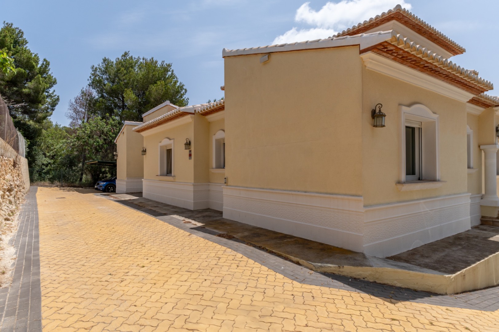 Fachada exterior de tonos crema con molduras blancas y tejado de teja cerámica. El camino de acceso está pavimentado con adoquines amarillos junto a una zona de aparcamiento cubierto.
